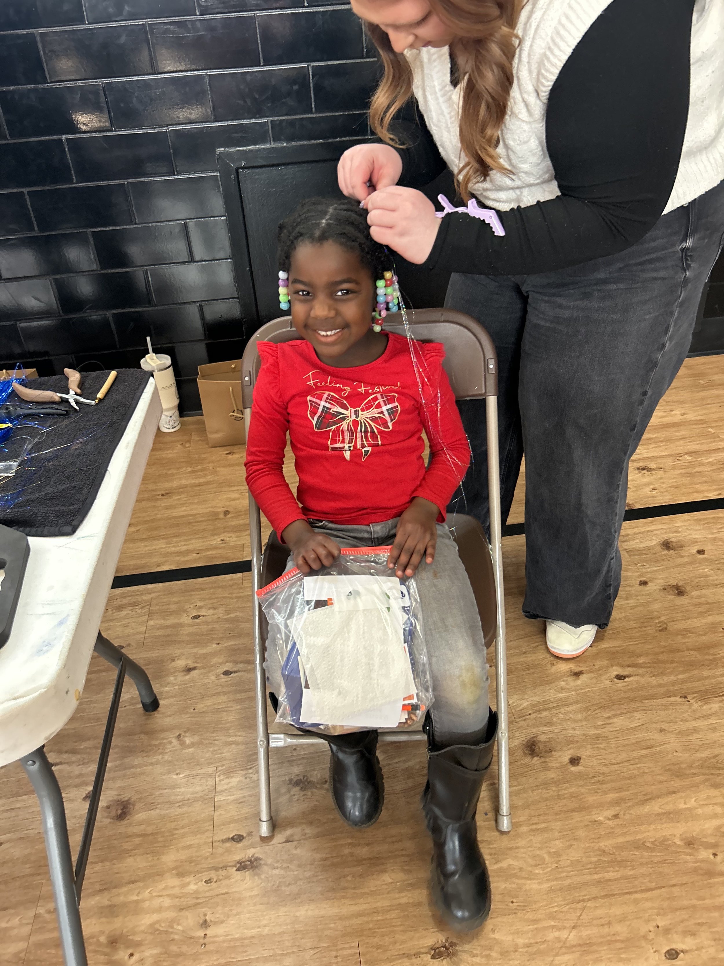 A young girl with colorful beaded hair accessories smiling while sitting on a chair, with a woman attaching her hair. The girl is wearing a red long-sleeve shirt with a bow graphic and some holiday writing, gray pants, and black boots. A plastic bag 
