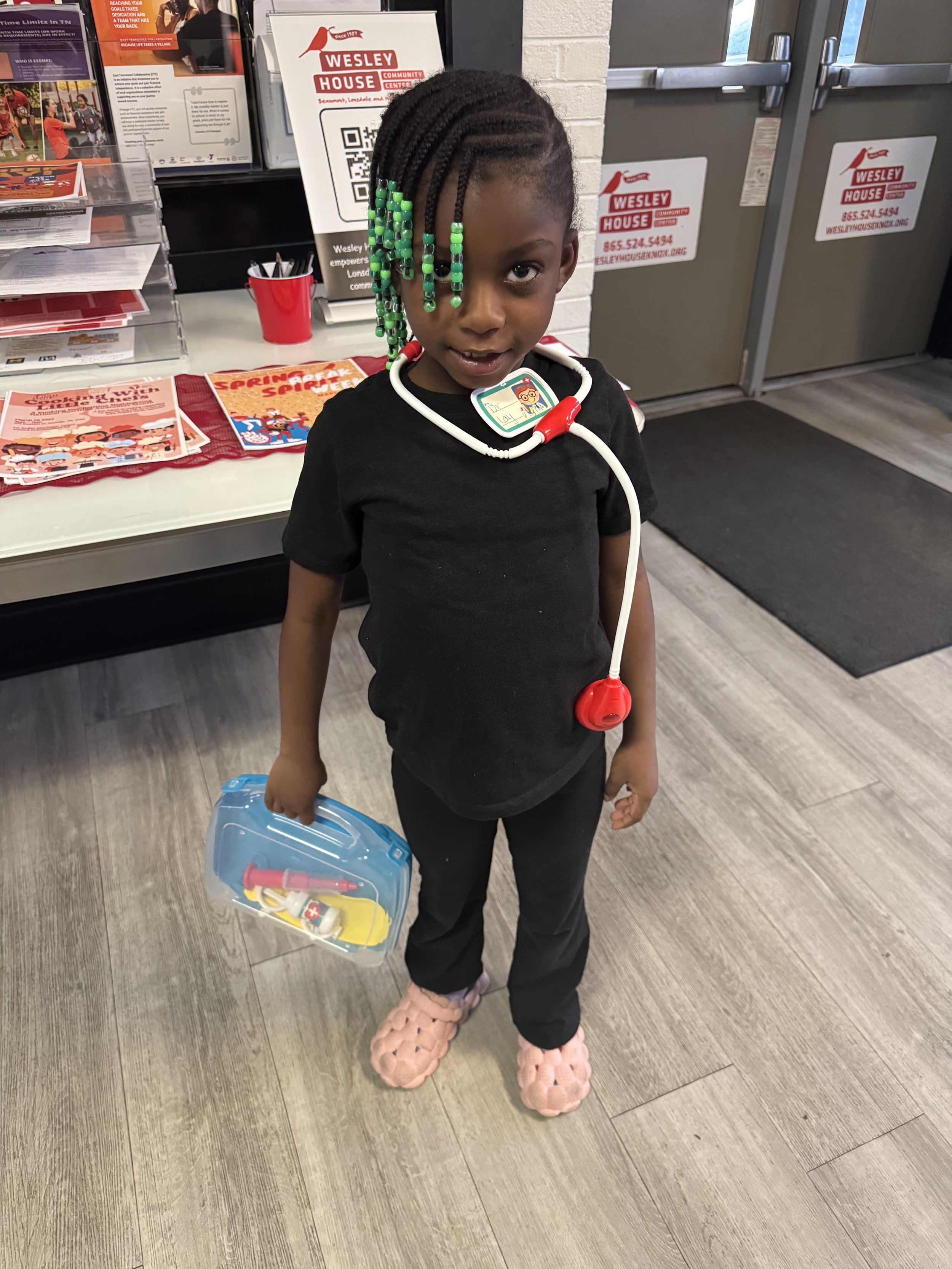 A young girl standing indoors, dressed as a doctor, wearing a toy stethoscope around her neck, holding a plastic medical toy case, and wearing pink fluffy slippers. She has braided hair with colorful beads and is smiling at the camera.