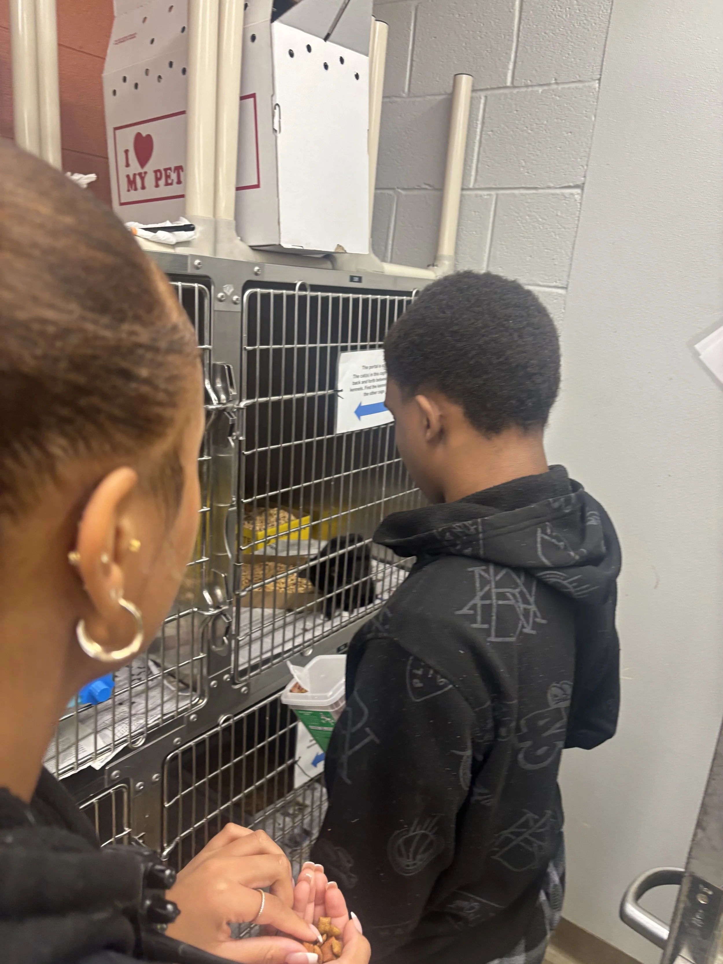 Two people, a woman and a young man, are at a pet store. The woman is holding pet food and looking at an animal inside a wire cage. The young man is facing the cage, likely watching or waiting.
