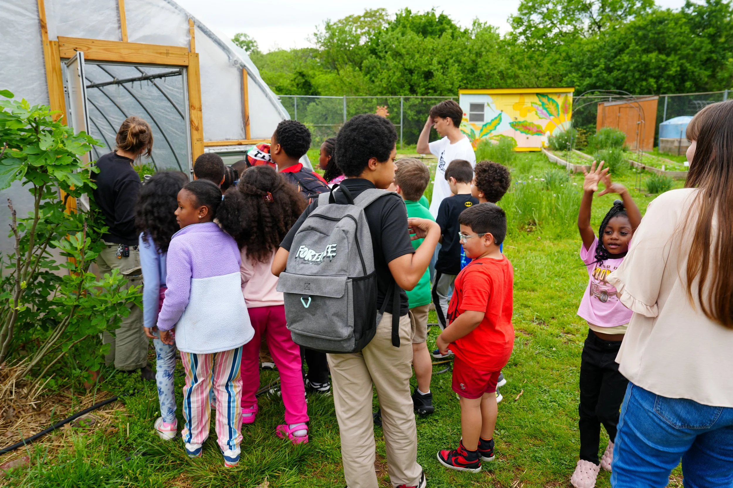 Group of children and two adults in a garden, with a greenhouse and a colorful mural on a small building in the background.