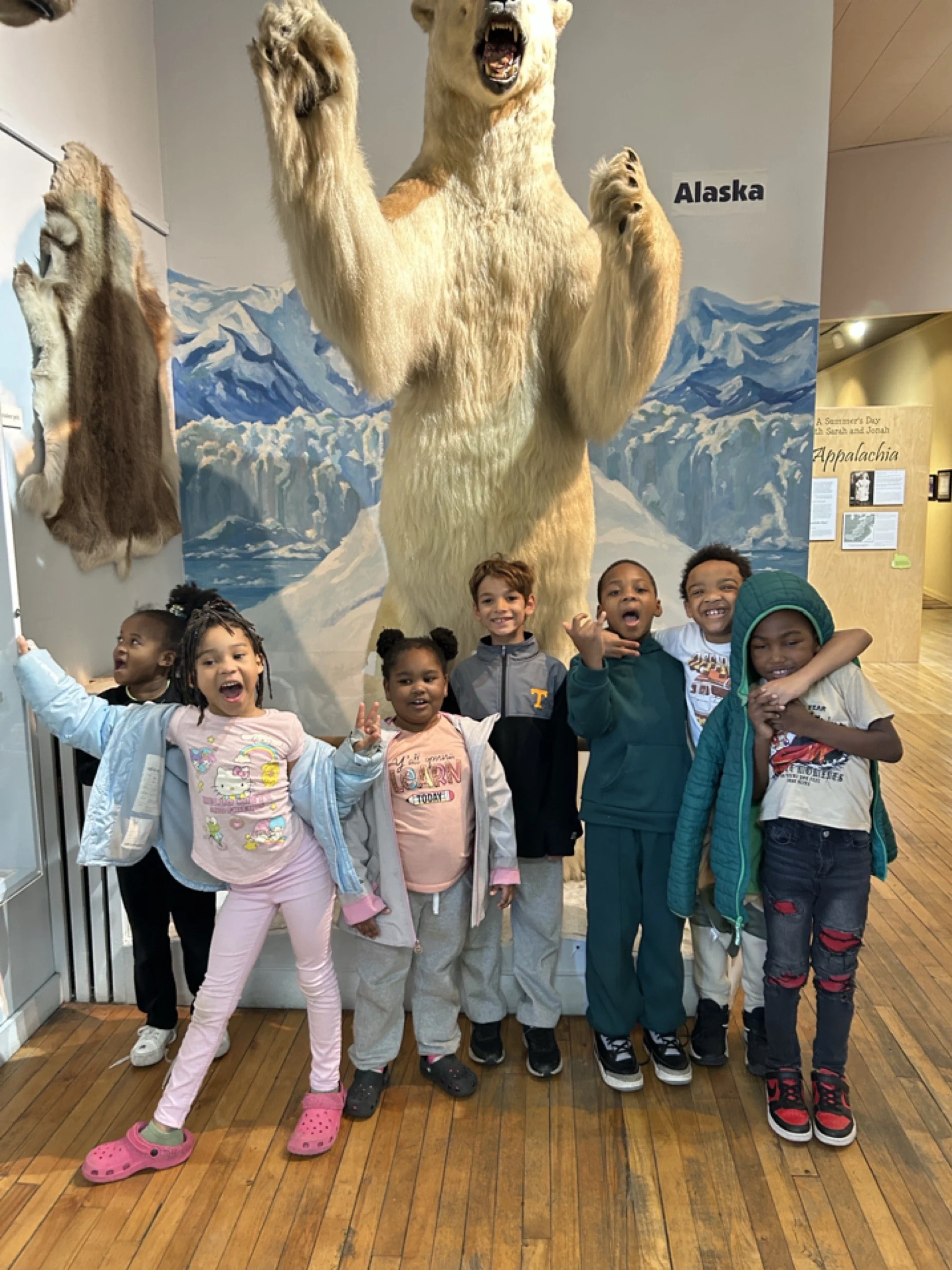 Group of seven children standing in front of a museum display of a large polar bear and its cub, with a mural of snowy mountains behind them. The children are smiling and posing for the photo.