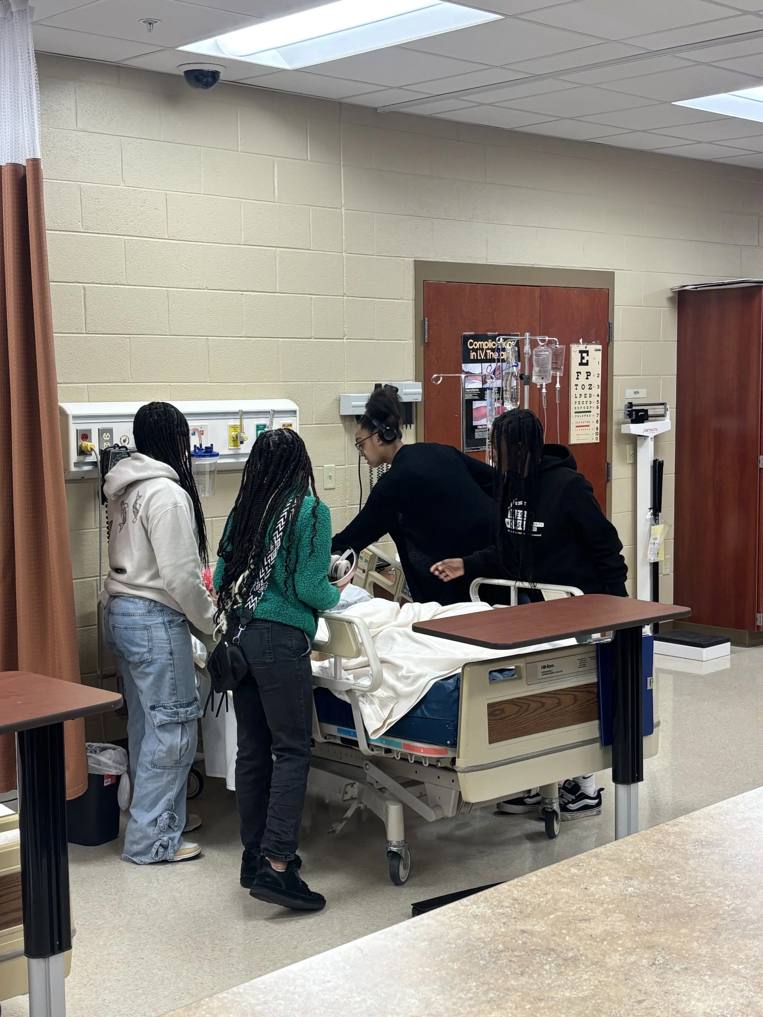 Four individuals are gathered around a hospital bed in a medical setting, with one person interacting with medical equipment. A patient lies in the bed covered with a white sheet, and IV stands are nearby.