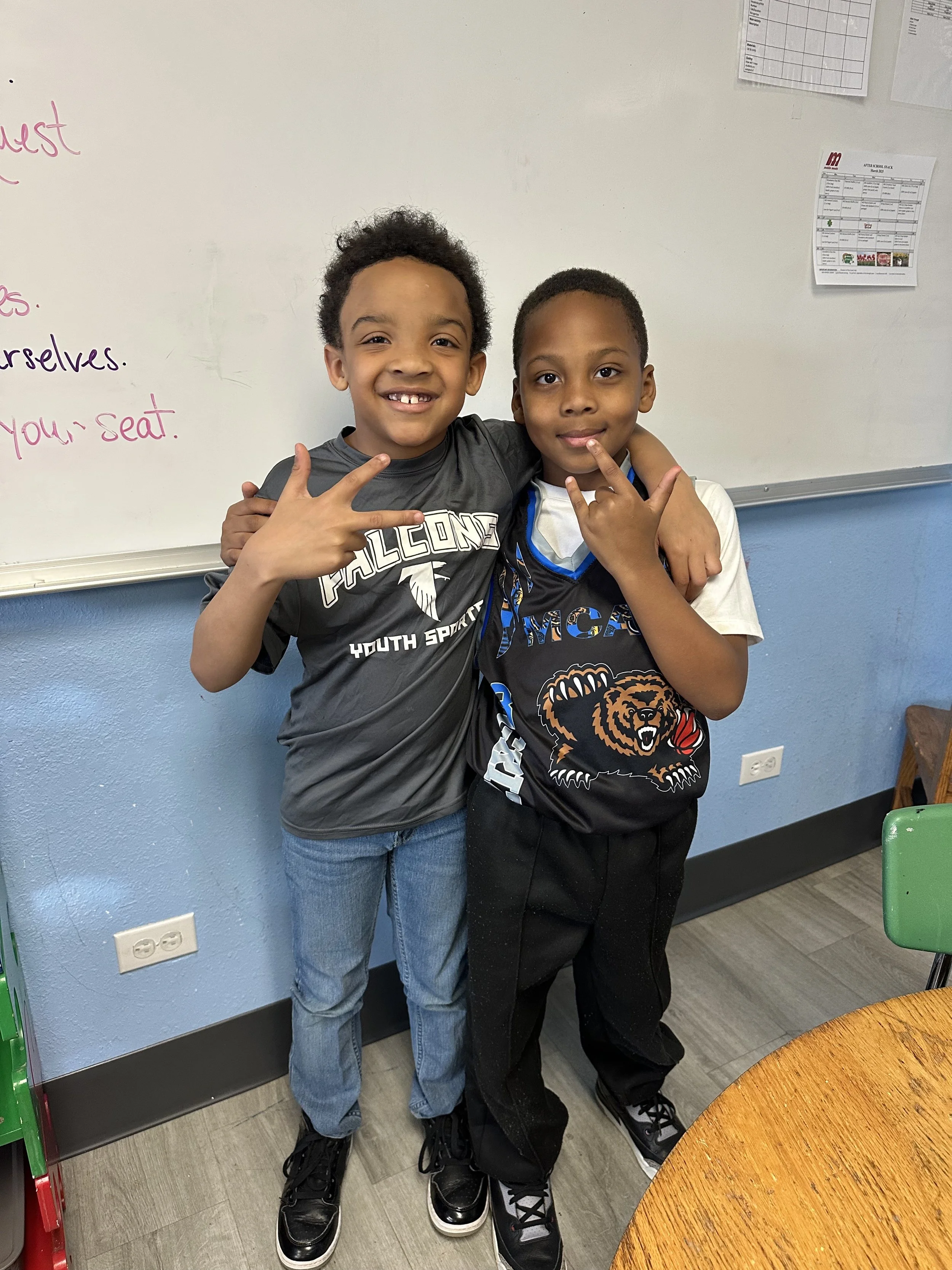 Two young boys standing near a classroom whiteboard, smiling and making hand signs. One boy is wearing a gray sports shirt with 'Falcons Youth Sports' and the other a black and blue jersey with a bear. The classroom has blue walls and a wooden table.