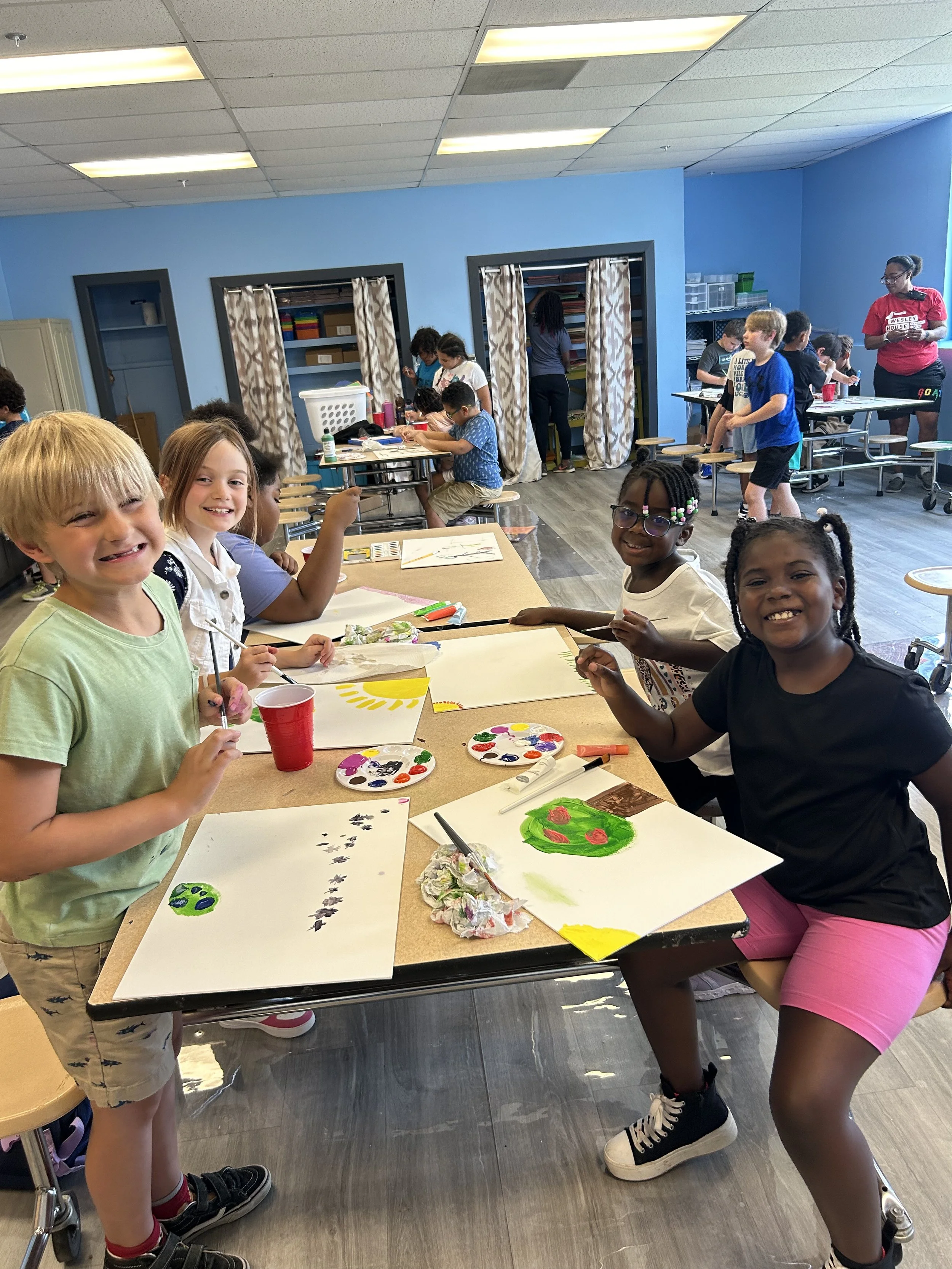 Children painting and drawing at a table in an art classroom with other students working in the background.