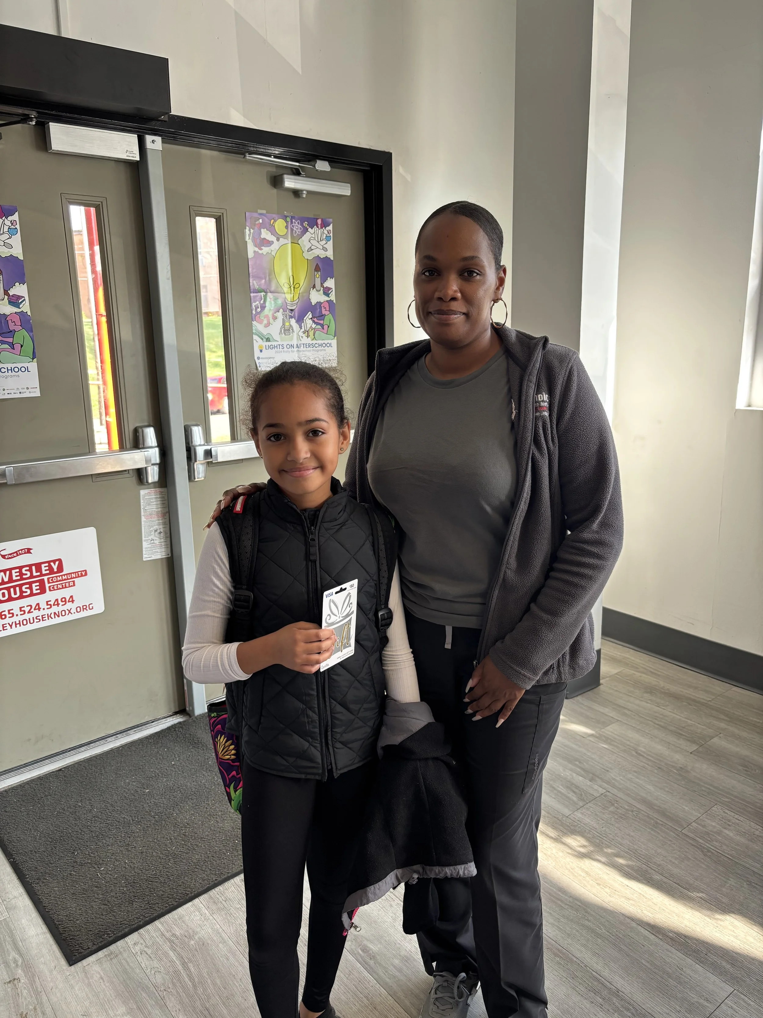 A woman and young girl standing together inside a building in front of a door, both smiling, with the girl holding a card. The girl has her hair tied back and wears a black vest, white long-sleeve shirt, and black pants. The woman wears a gray shirt,
