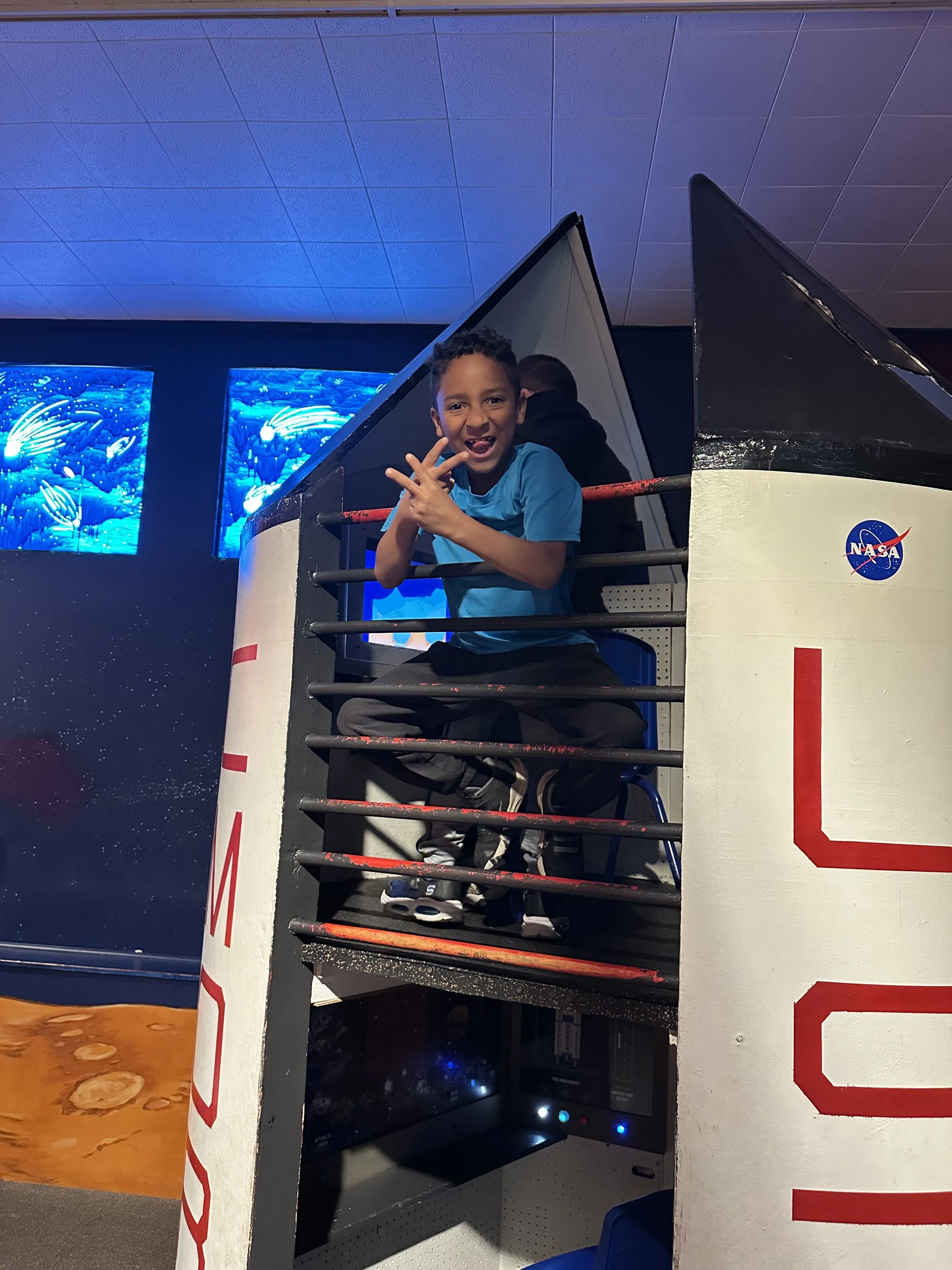 Child sitting on a NASA space shuttle replica in an indoor exhibit, smiling and making a hand gesture with a space-themed background.