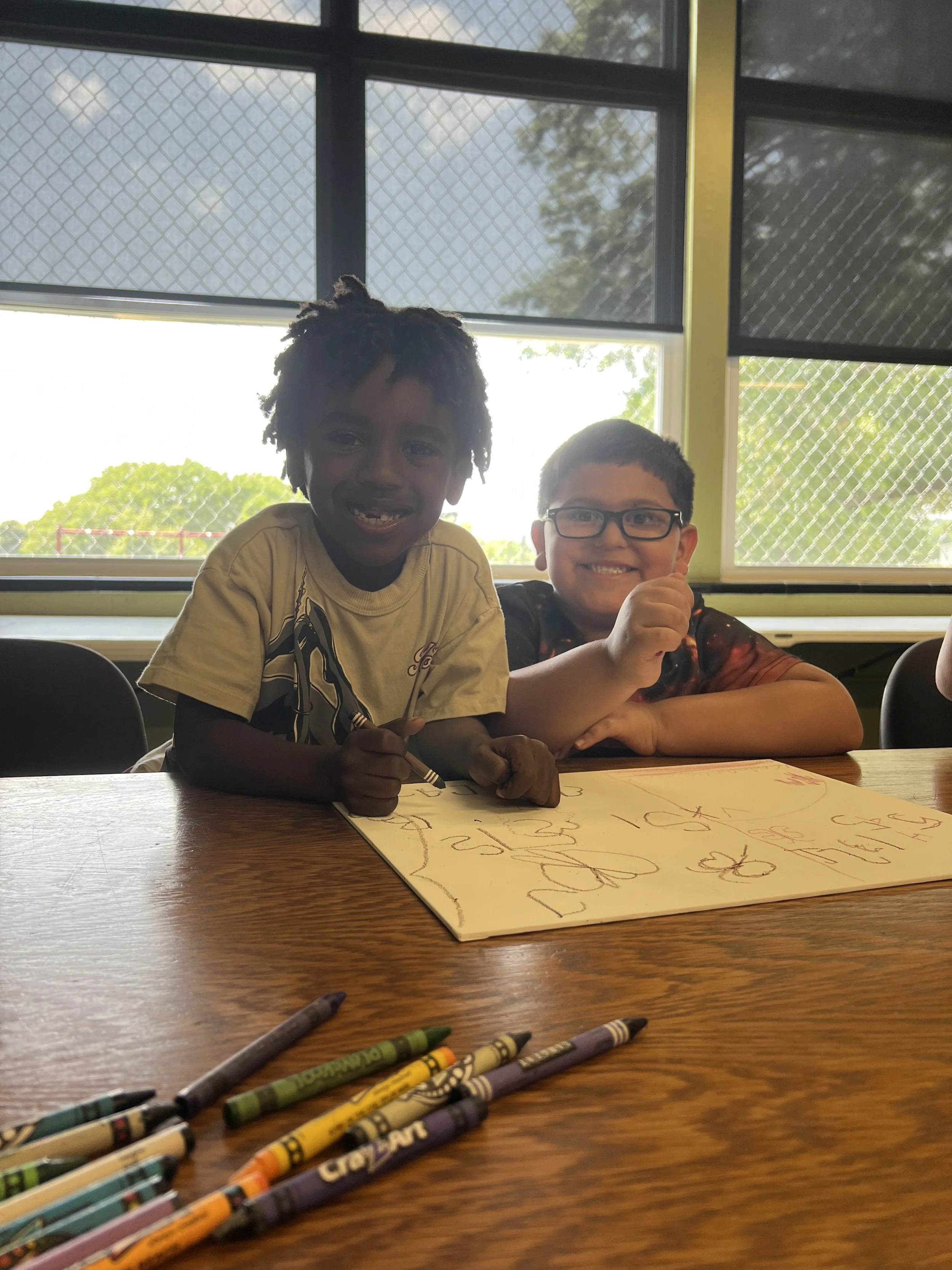 Two children, one with curly dark hair and glasses, smiling, sitting at a table with crayons and a drawing, in front of a window with a mesh screen, bright outdoors.