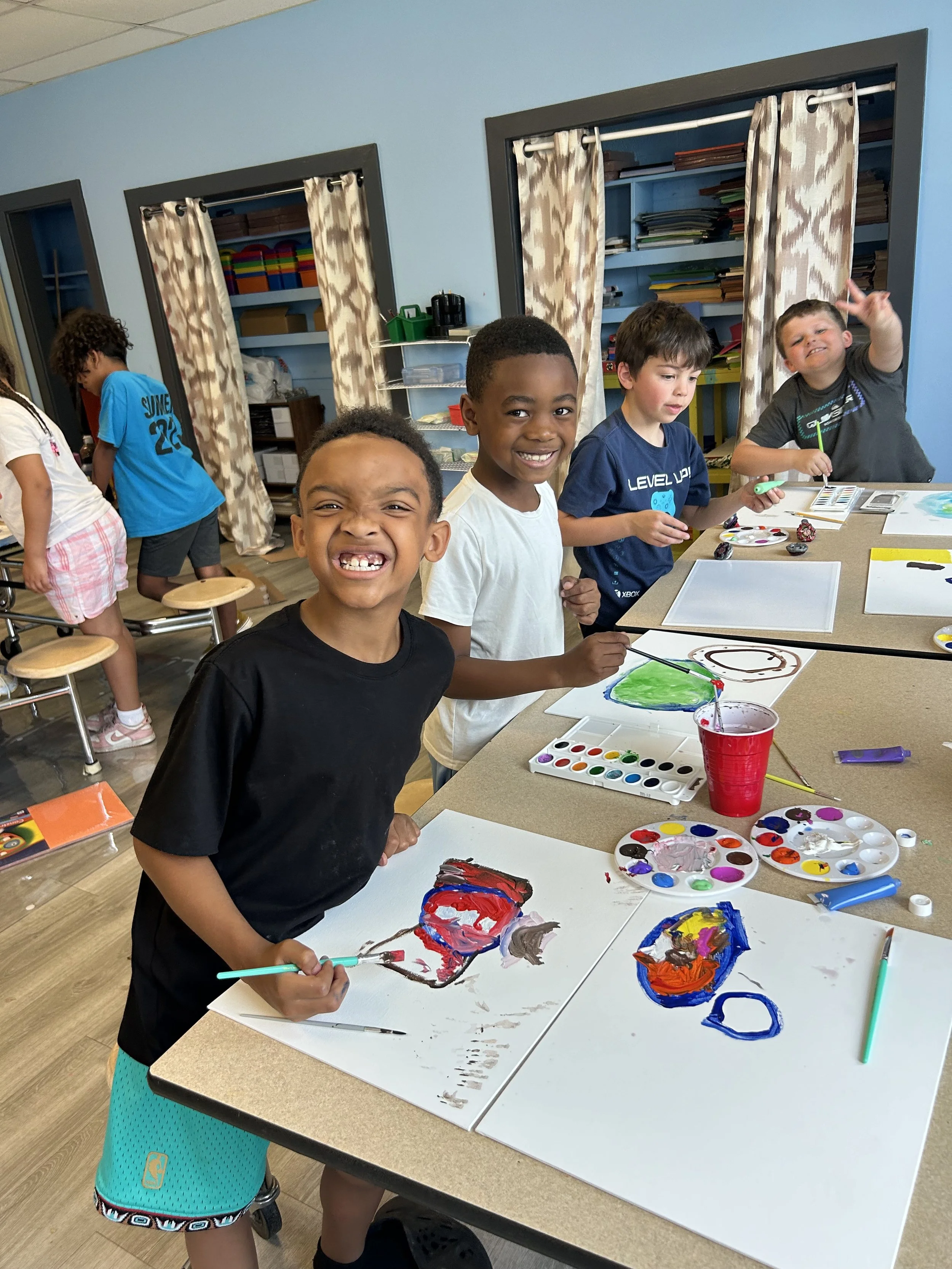 Children in a classroom painting and smiling with paintings on the table.