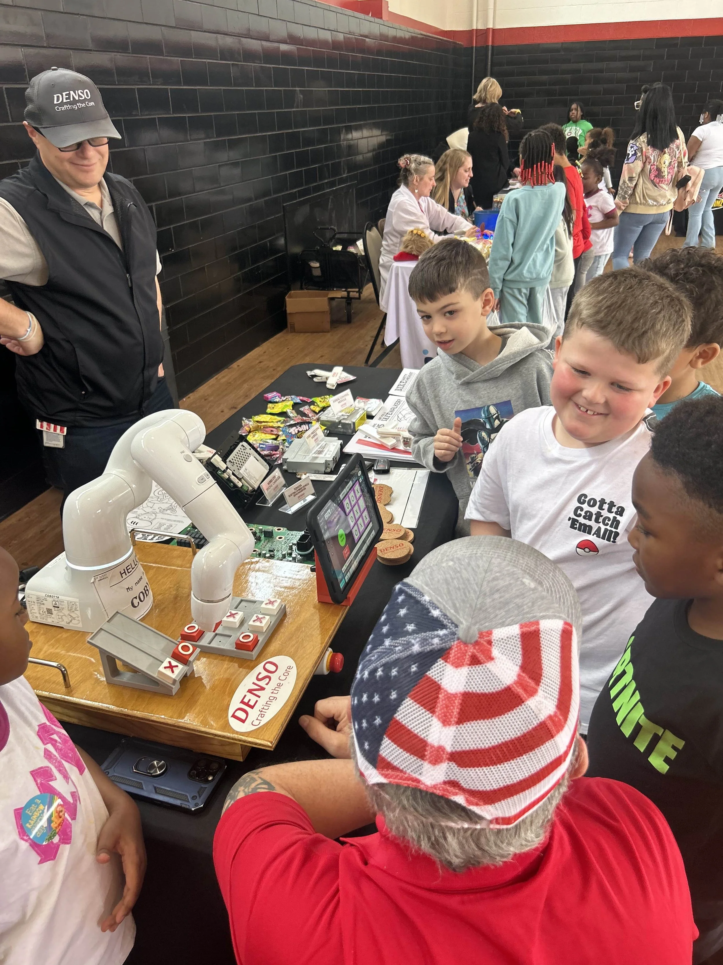 Children gathered around a demonstration table at a science or robotics event, with a person operating a robotic arm.