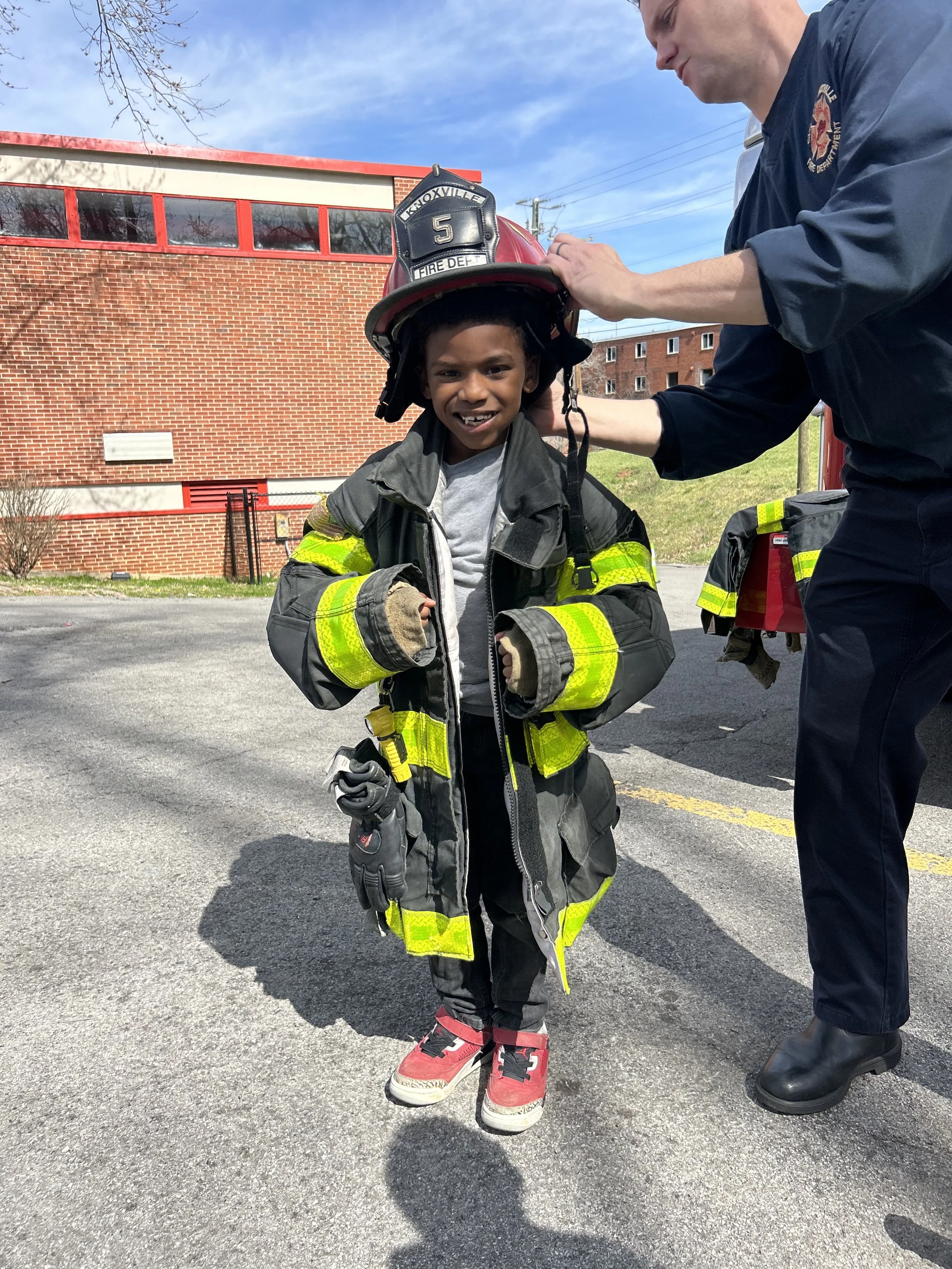 A young boy wearing a vintage firefighter's coat and boots, smiling while being fitted for a firefighter's helmet by an adult firefighter outside in a parking lot with a brick building in the background.
