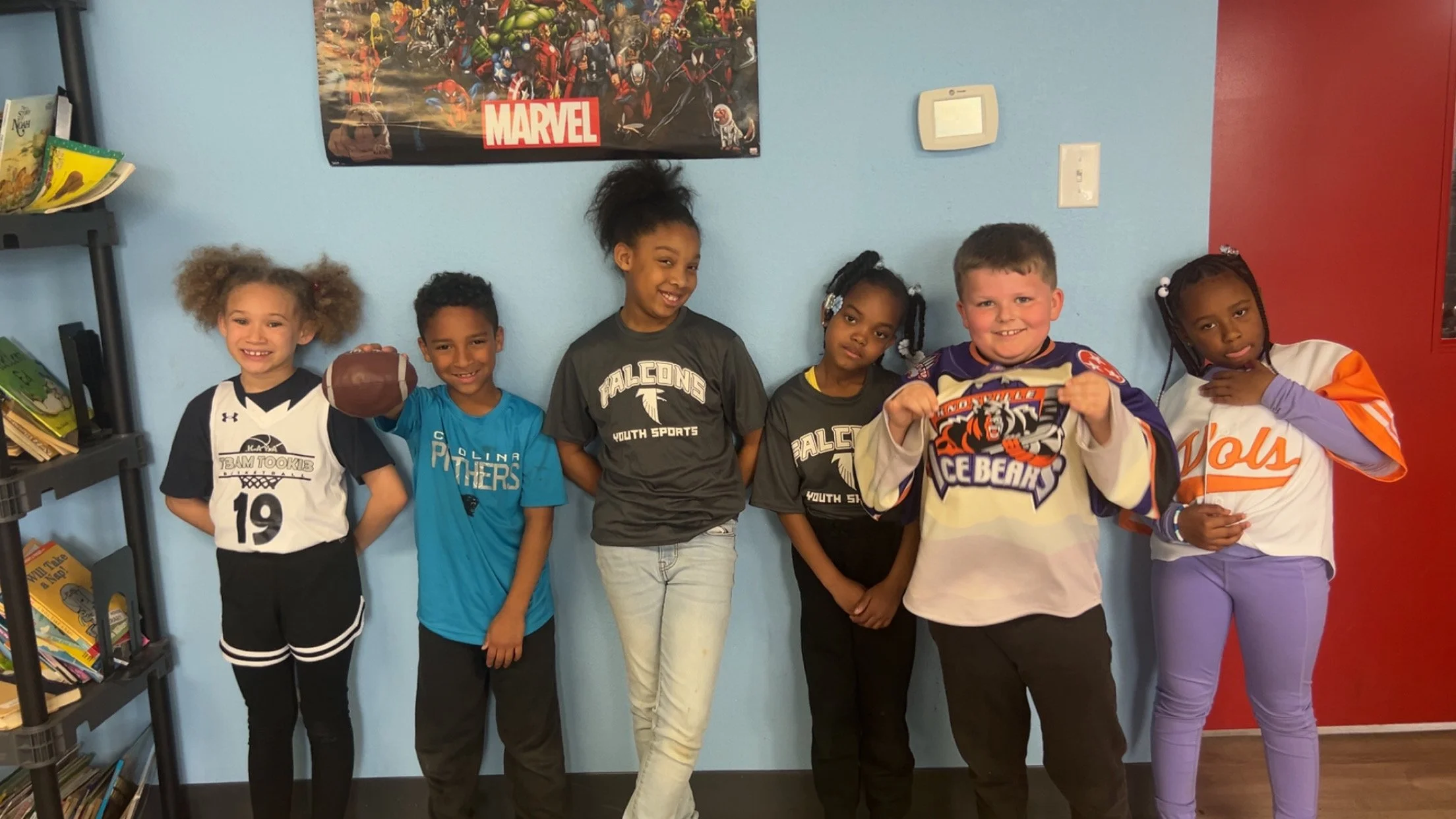 Six children standing against a blue wall with a Marvel poster. The children are smiling or making playful faces, dressed in sports jerseys or casual clothing, with one holding a football.