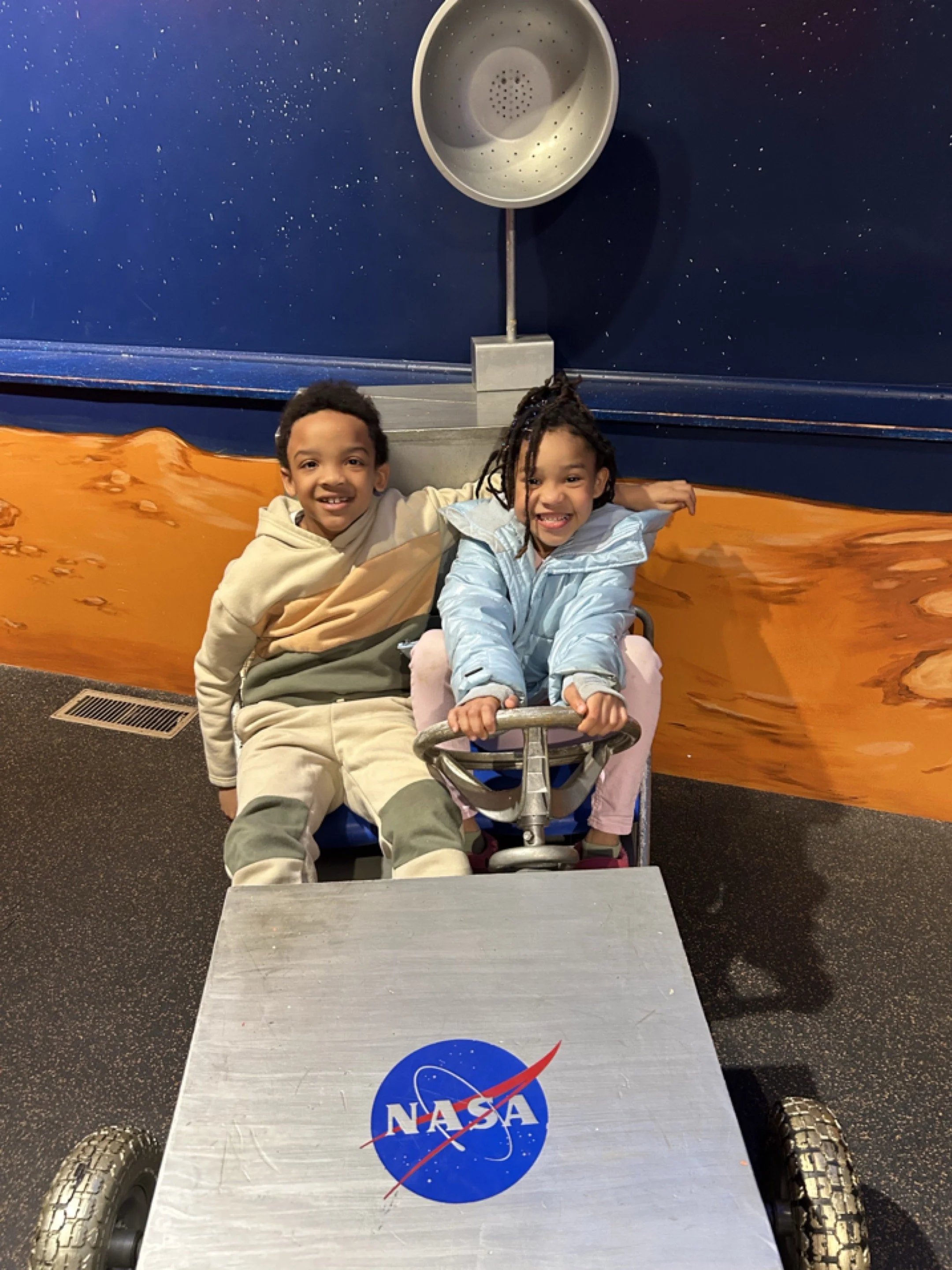 Two children sitting in a NASA-themed space exhibit, smiling at the camera, with a space-themed mural and a showerhead-like object behind them.