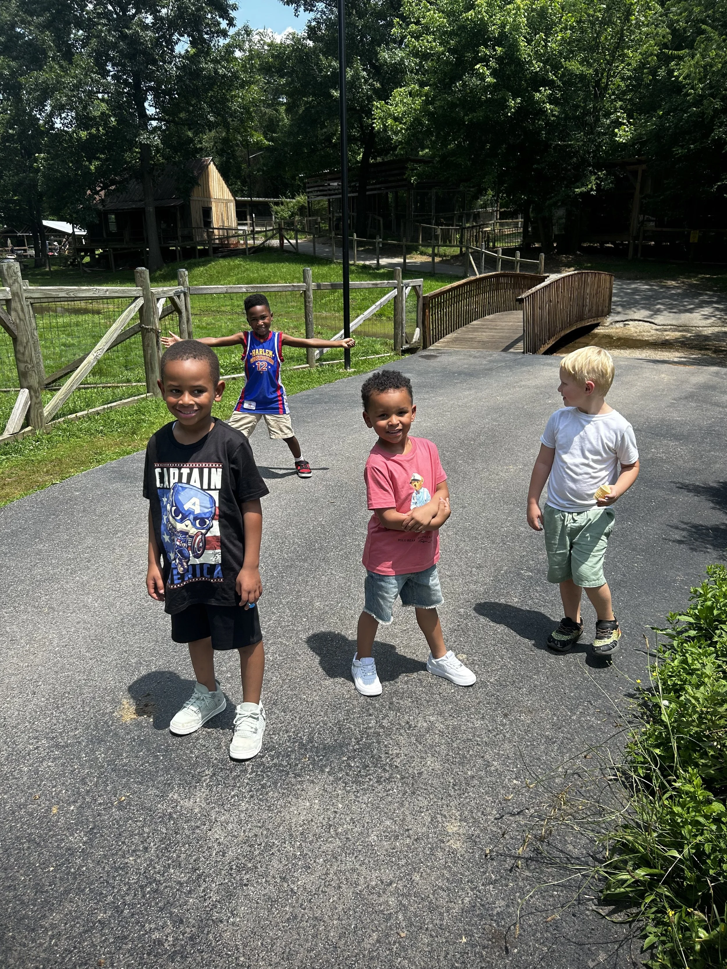 Four young children standing outdoors on a paved path at a park with wooden fencing, trees, and small wooden structures in the background. One child is in the background with arms outstretched. The children are smiling and dressed casually.