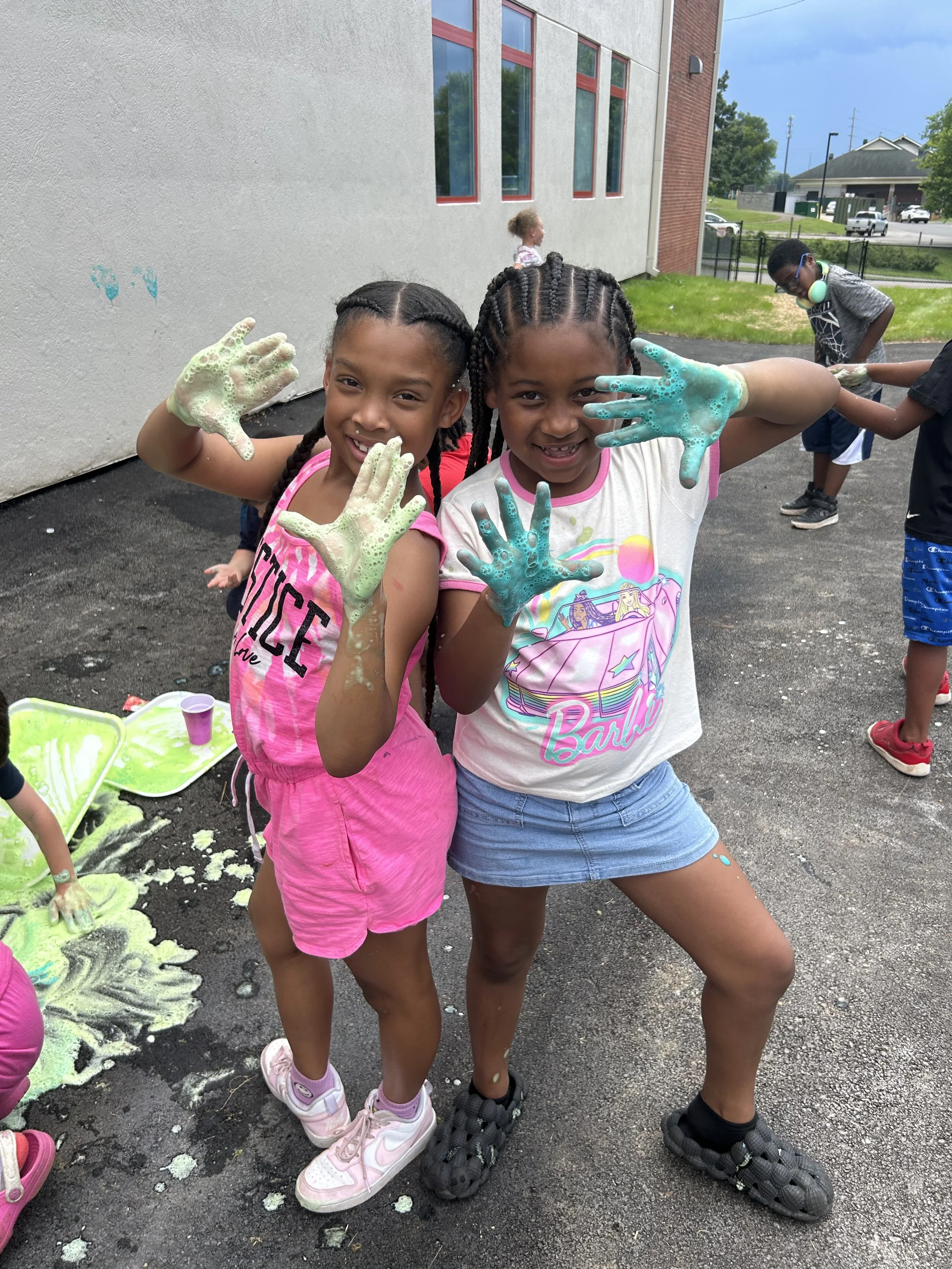 Two young girls with braided hair and colorful shirts are smiling and showing their soapy hands after playing with foam on a sunny day outdoors.