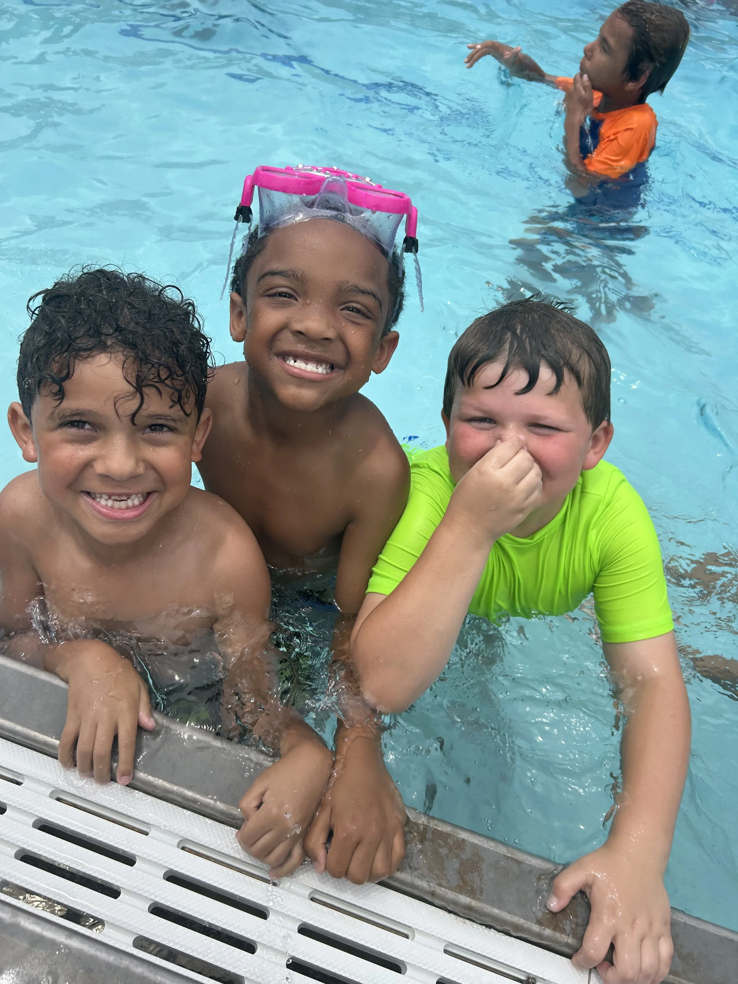 Three kids smiling and enjoying in a swimming pool, with one wearing goggles, and another pinching his nose, while a fourth person is swimming in the background.