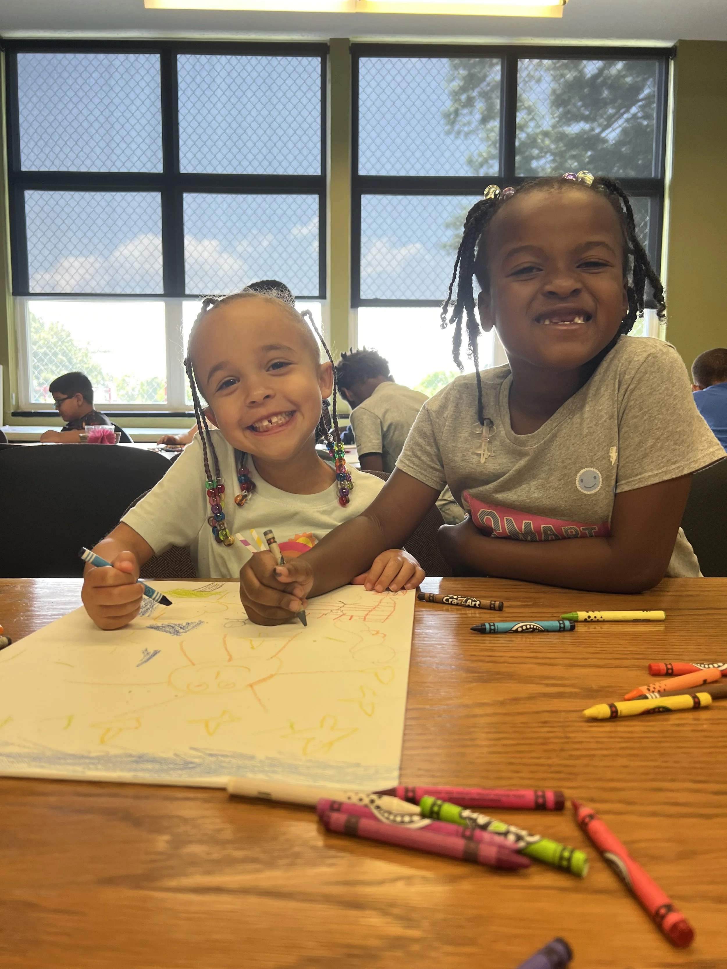 Two young girls smiling and drawing on a large sheet of paper with colorful crayons in an indoor classroom setting, with windows showing a partly cloudy sky outside.