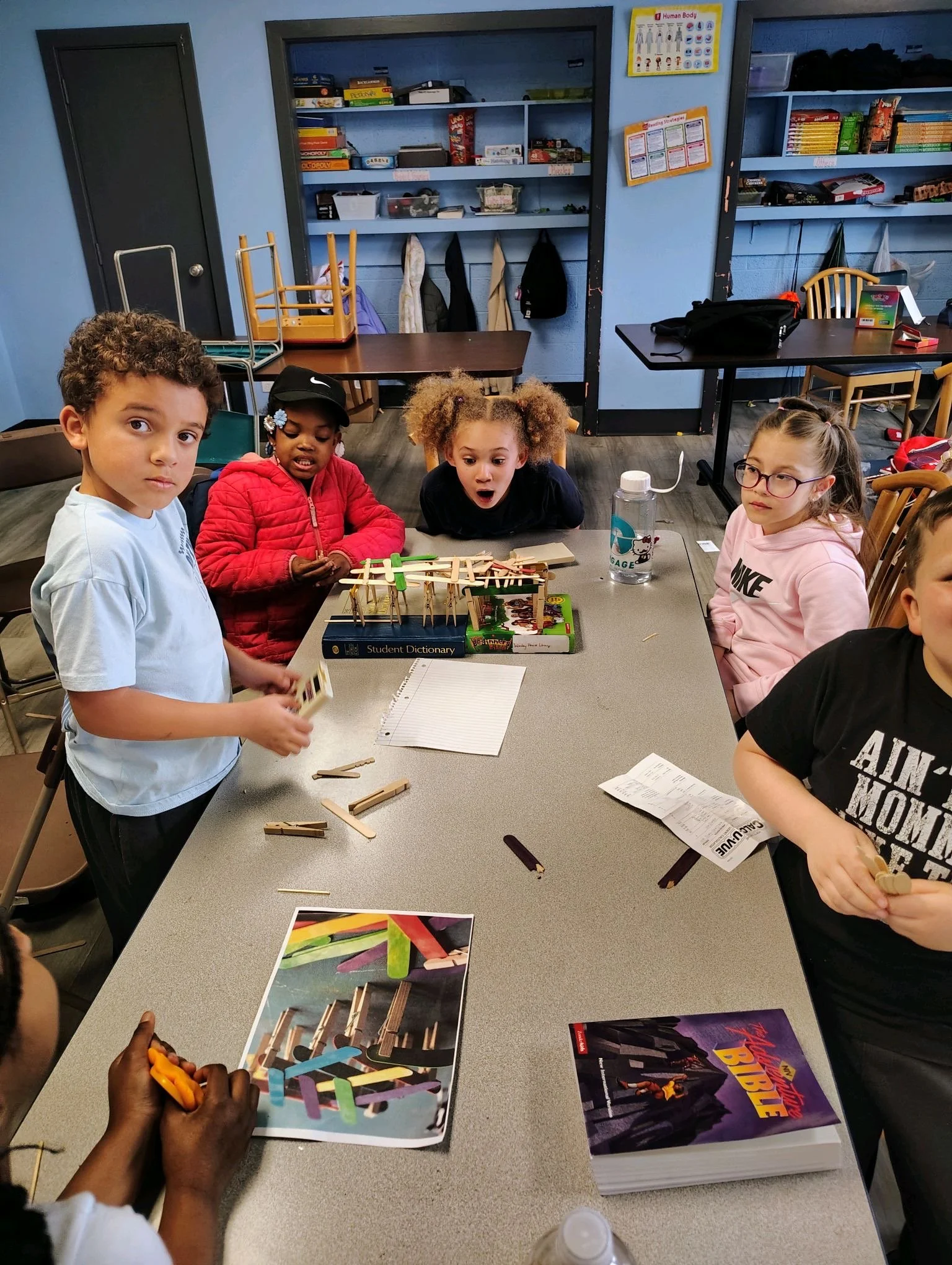 Children gathered around a table working on a craft project with popsicle sticks, glue, and marker; a colorful illustration and a Bible are on the table; classroom setting with educational posters, bookshelves, and jackets hanging on hooks in the bac