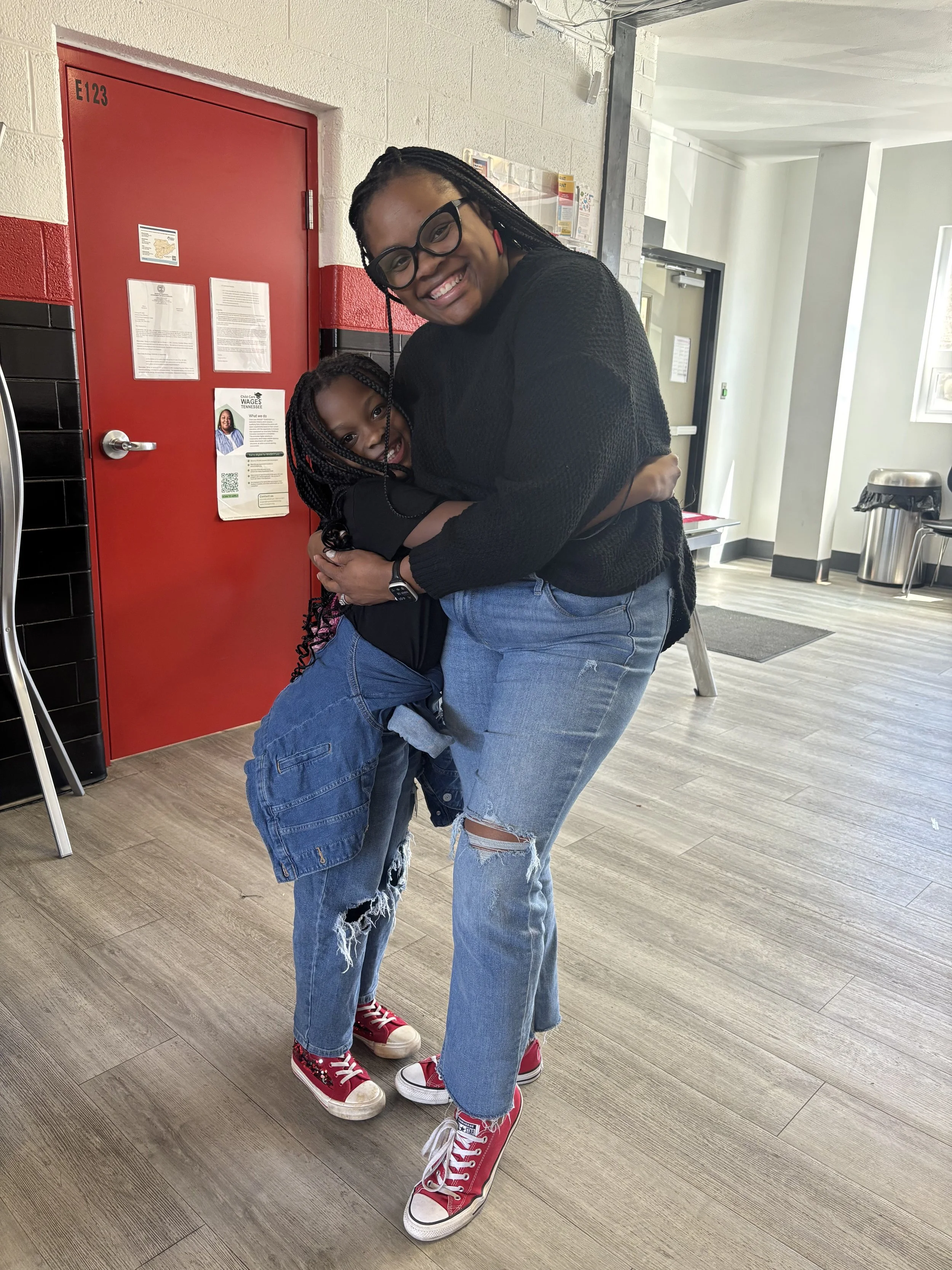 A woman and a girl hugging and smiling inside a building, with a red door and wooden flooring.