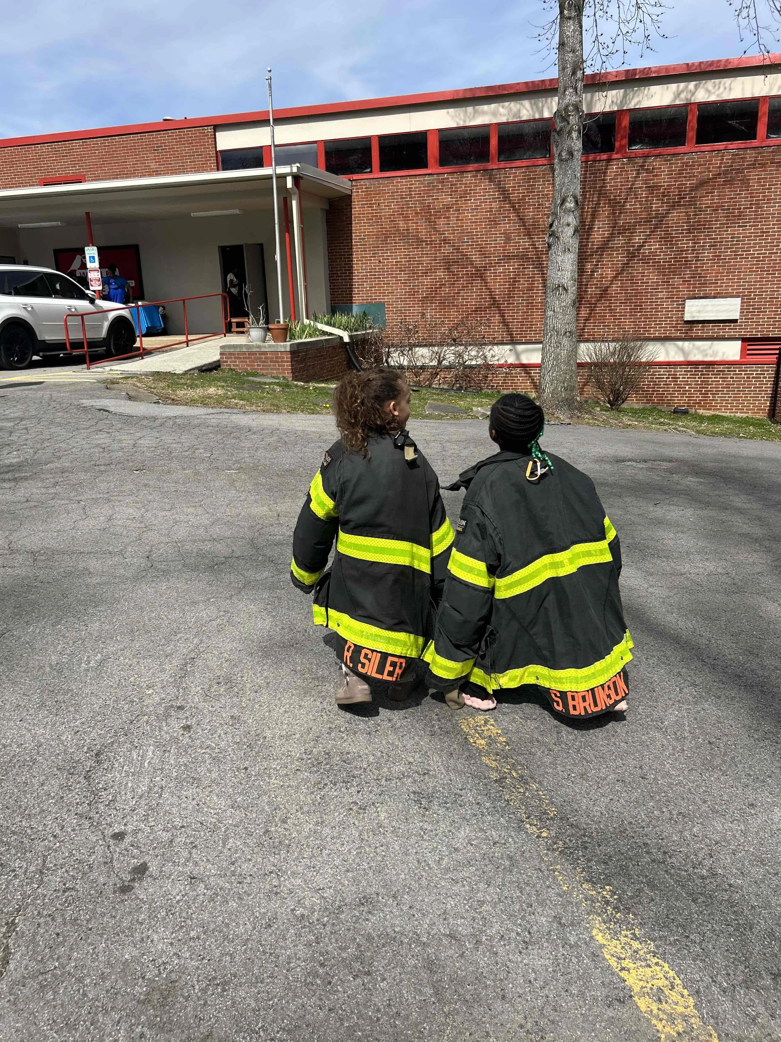 Two children dressed as firefighters kneeling on a parking lot near a fire station, with a brick building, a parked car, and a tree in the background.