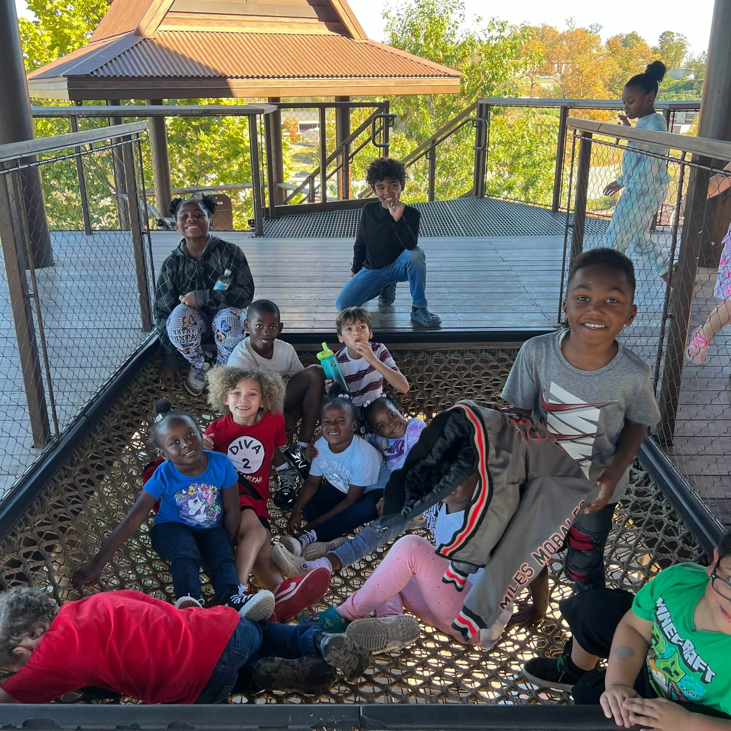 Children playing and sitting in a treehouse with a wooden floor and metal railings, surrounded by trees and a clear sky.