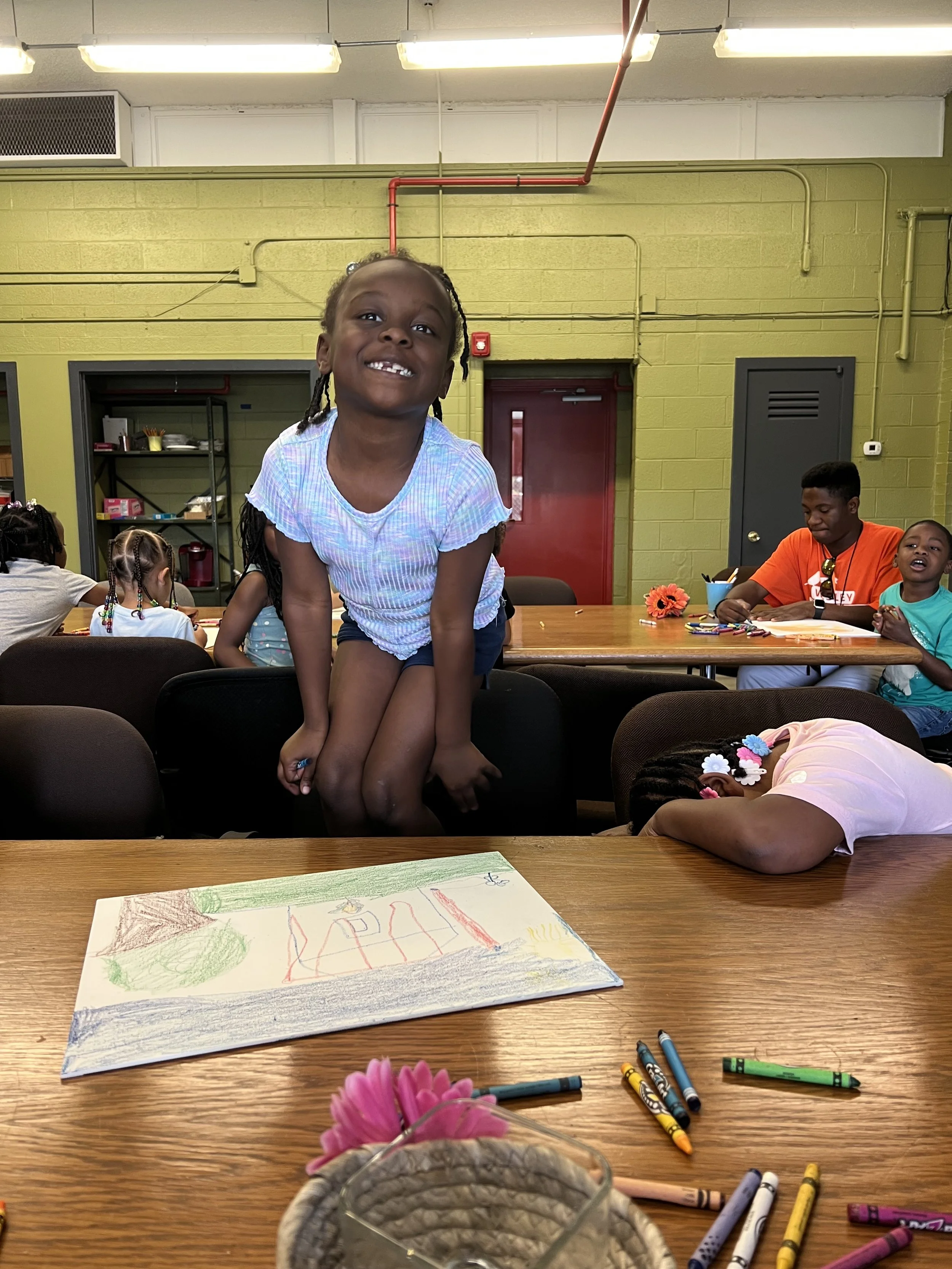 A young girl is jumping onto a chair in a classroom with other children and adults around her, colorful drawings and crayons on the table, green painted walls, and a red door in the background.