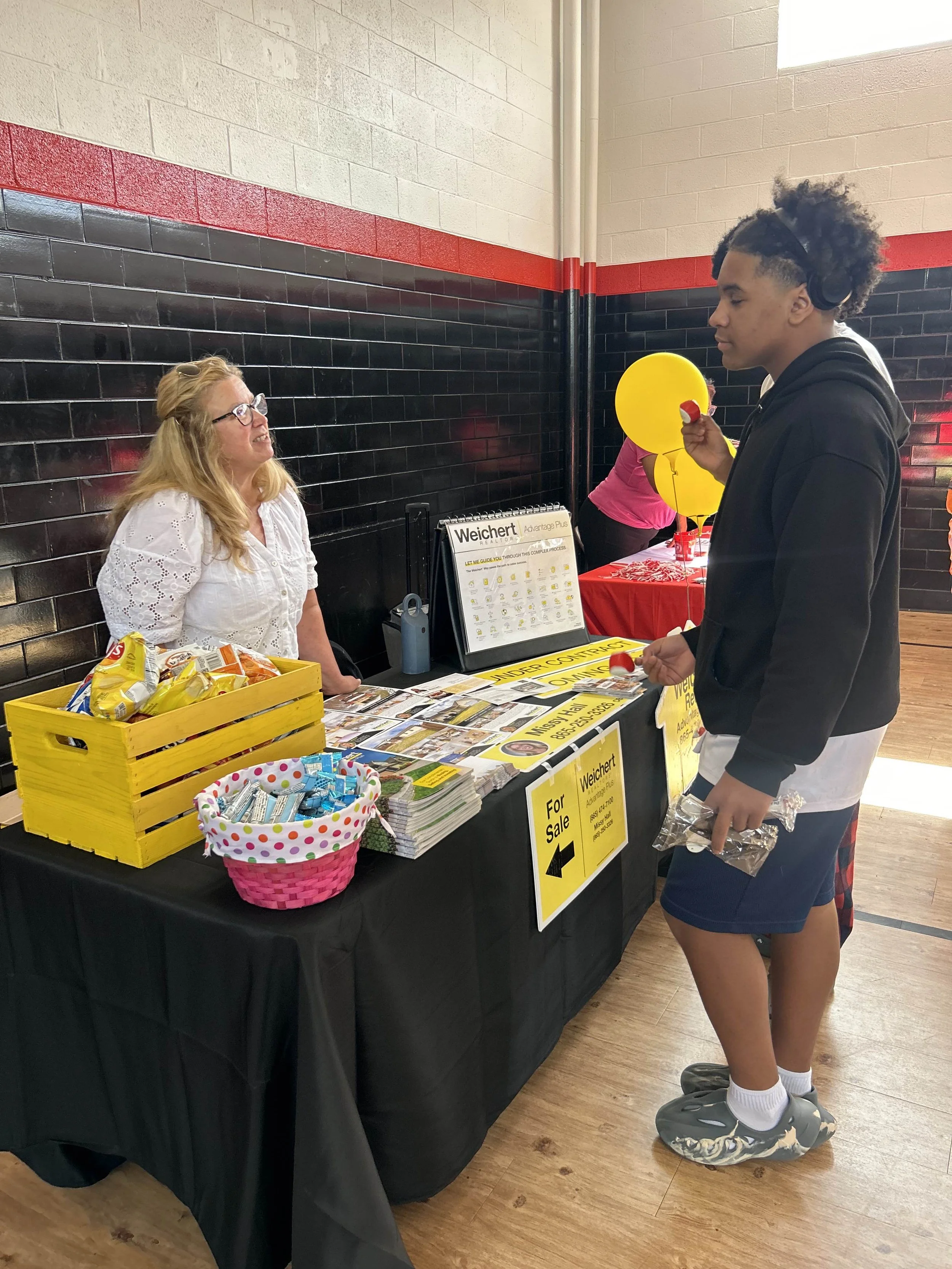 A young man shopping at a sale booth interacts with a woman behind the table, which displays various items for sale, including snacks and flyers, with a sign indicating "For Sale" and an arrow. The booth is decorated with yellow signs and balloons.