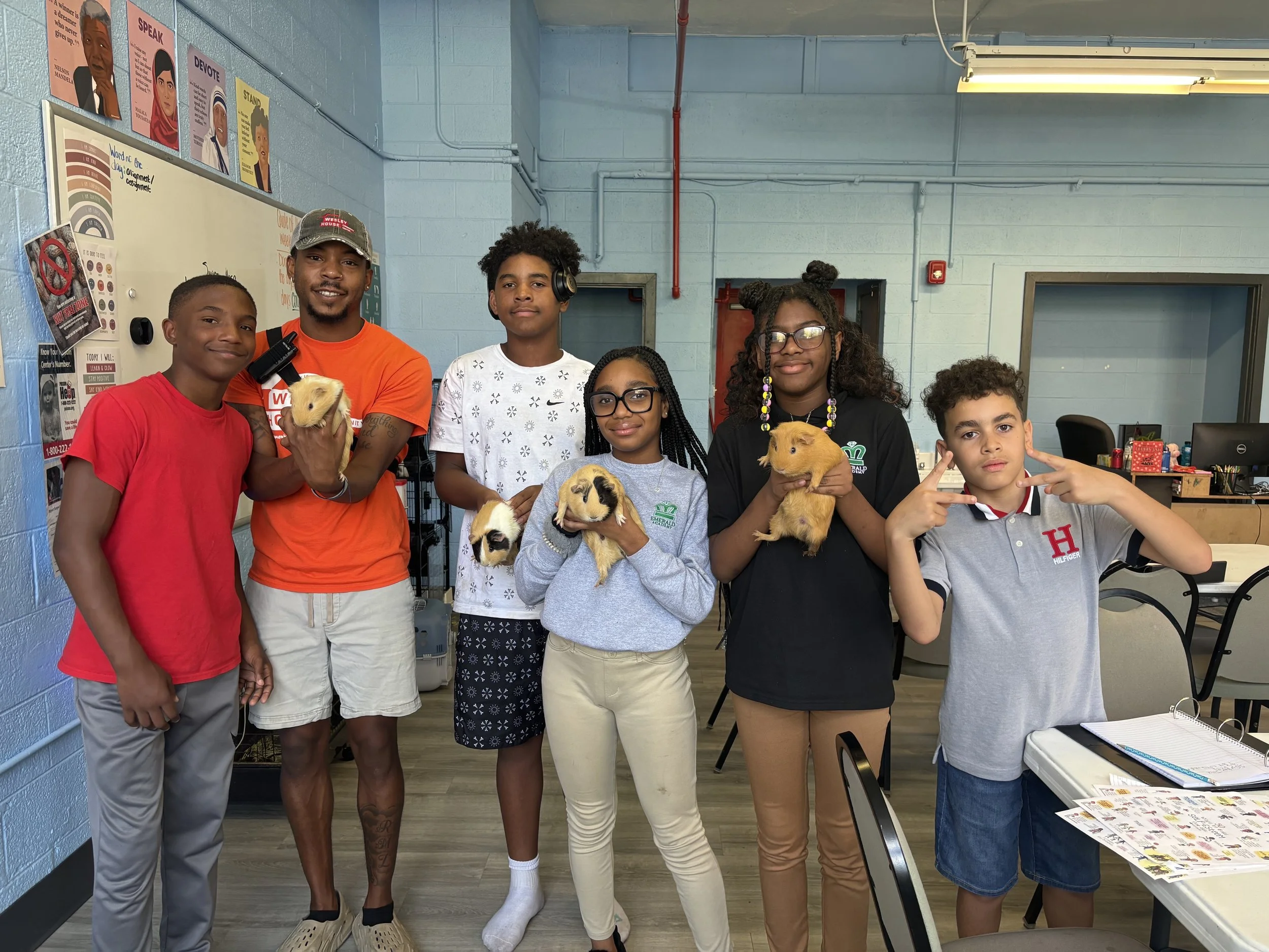 Group of seven diverse children and teenagers holding guinea pigs indoors, with classroom or community space background.