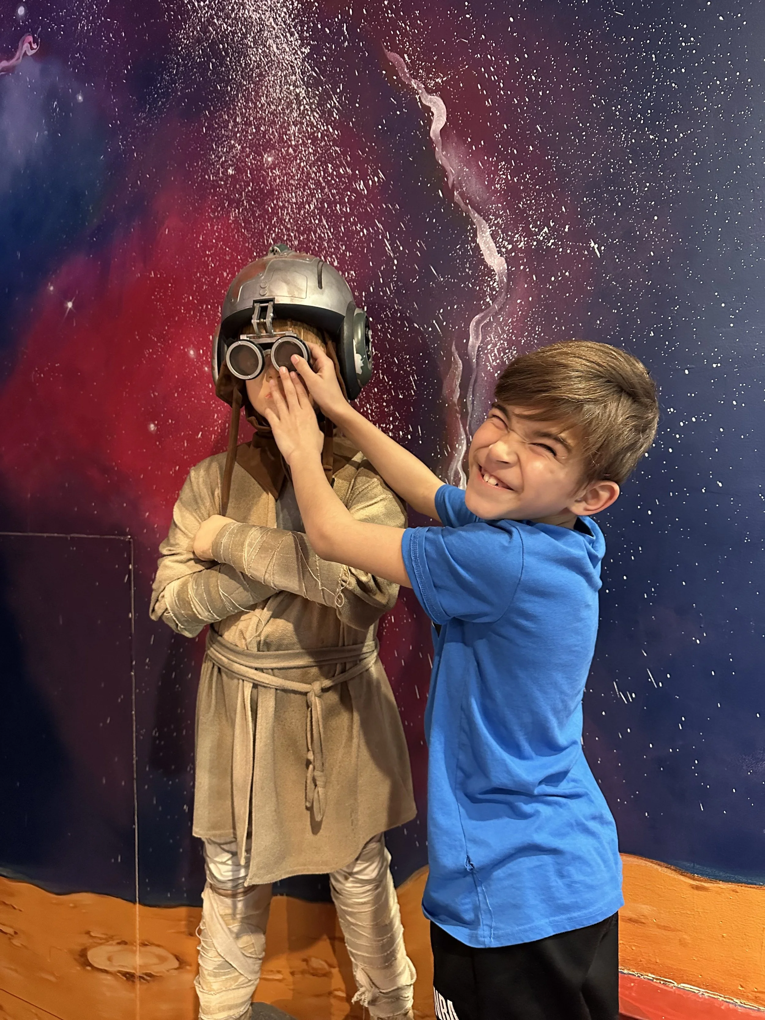 A young boy playfully covers a person dressed as a space explorer with a helmet and binoculars, standing in front of a colorful galaxy-themed backdrop.