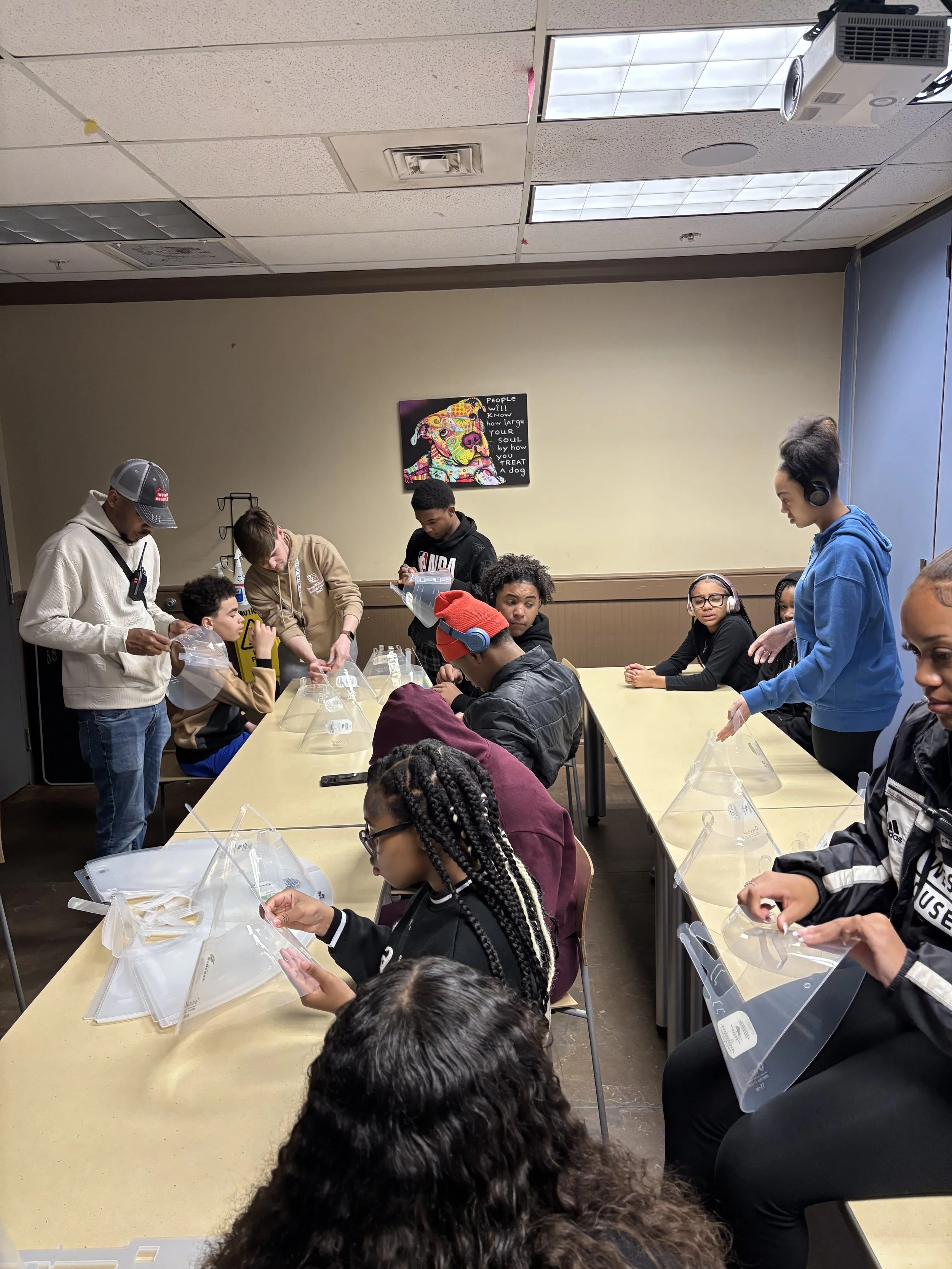 Group of children and teenagers sitting at tables with face shields, engaging in a hands-on activity in a classroom or meeting room.