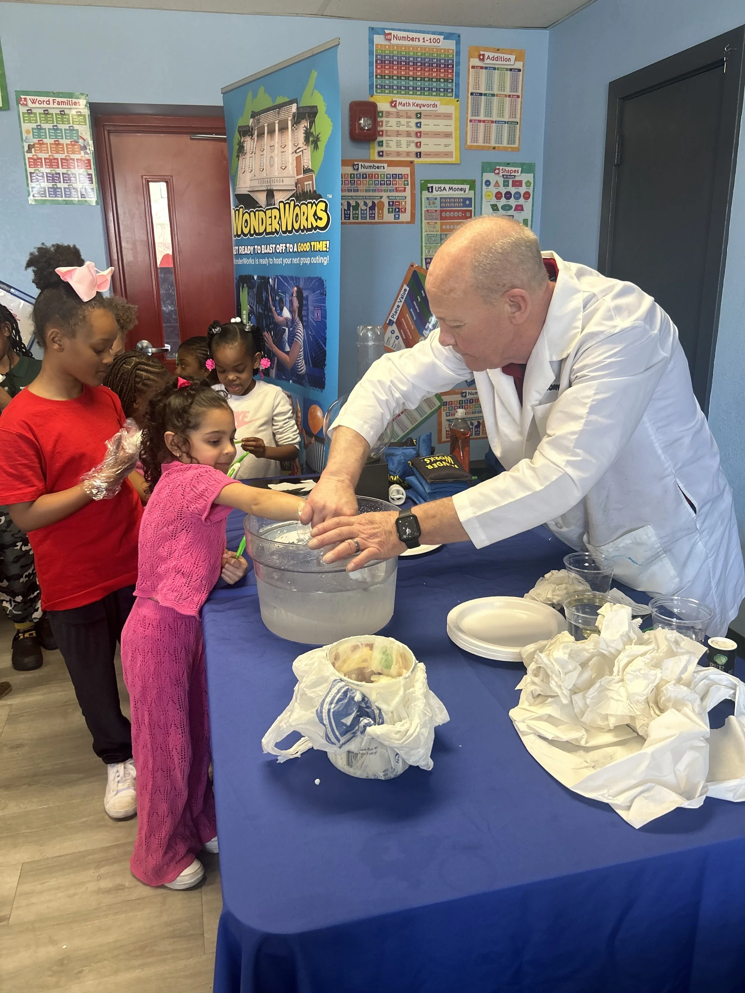 A man in a white lab coat performing a science experiment with children gathered around. Children are watching and touching the experiment, with posters and educational charts in the background.