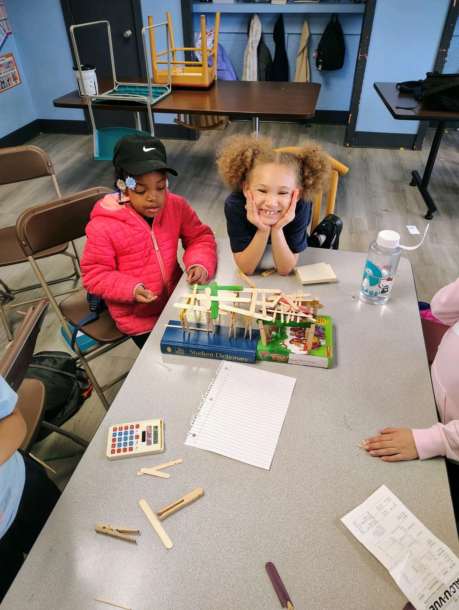 Two young girls at a table in a classroom, working on a wooden stick construction project, with a notebook, calculator, and water bottle on the table.