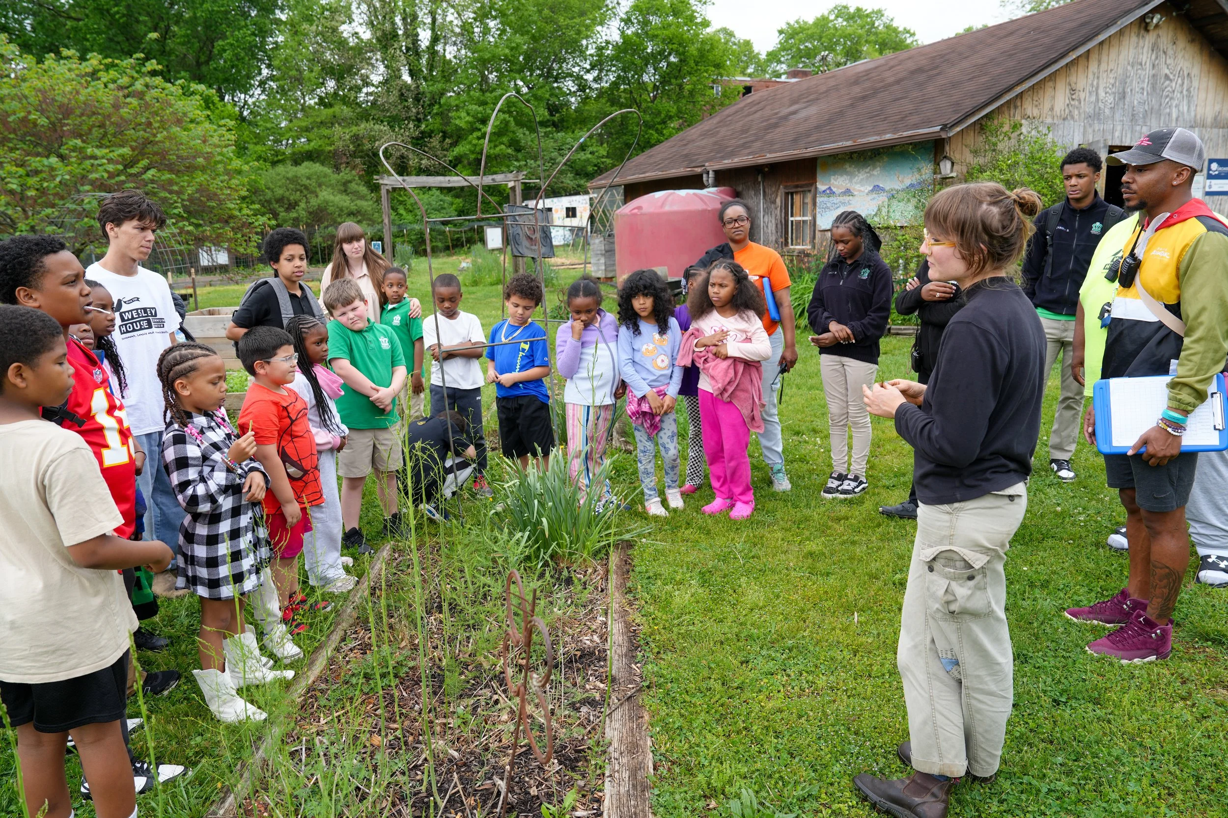 Group of children and adults gathered outdoors around a garden bed, listening to a woman speaking, in a rural setting with trees, a wooden shed, and a mural in the background.