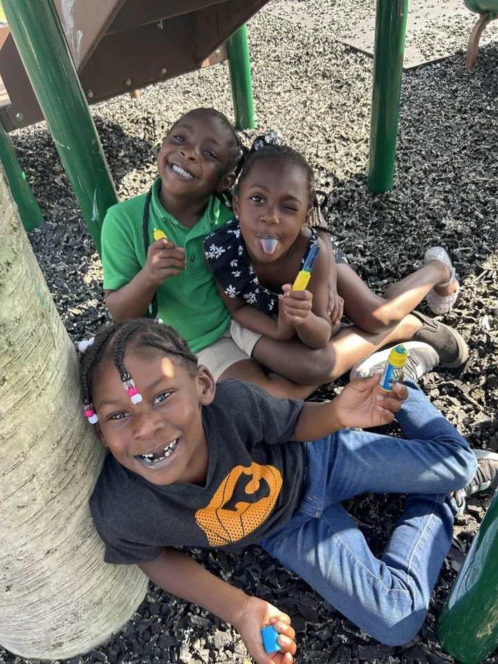 Three children sitting under a playground structure, smiling and holding bubble toys on a gravel surface.