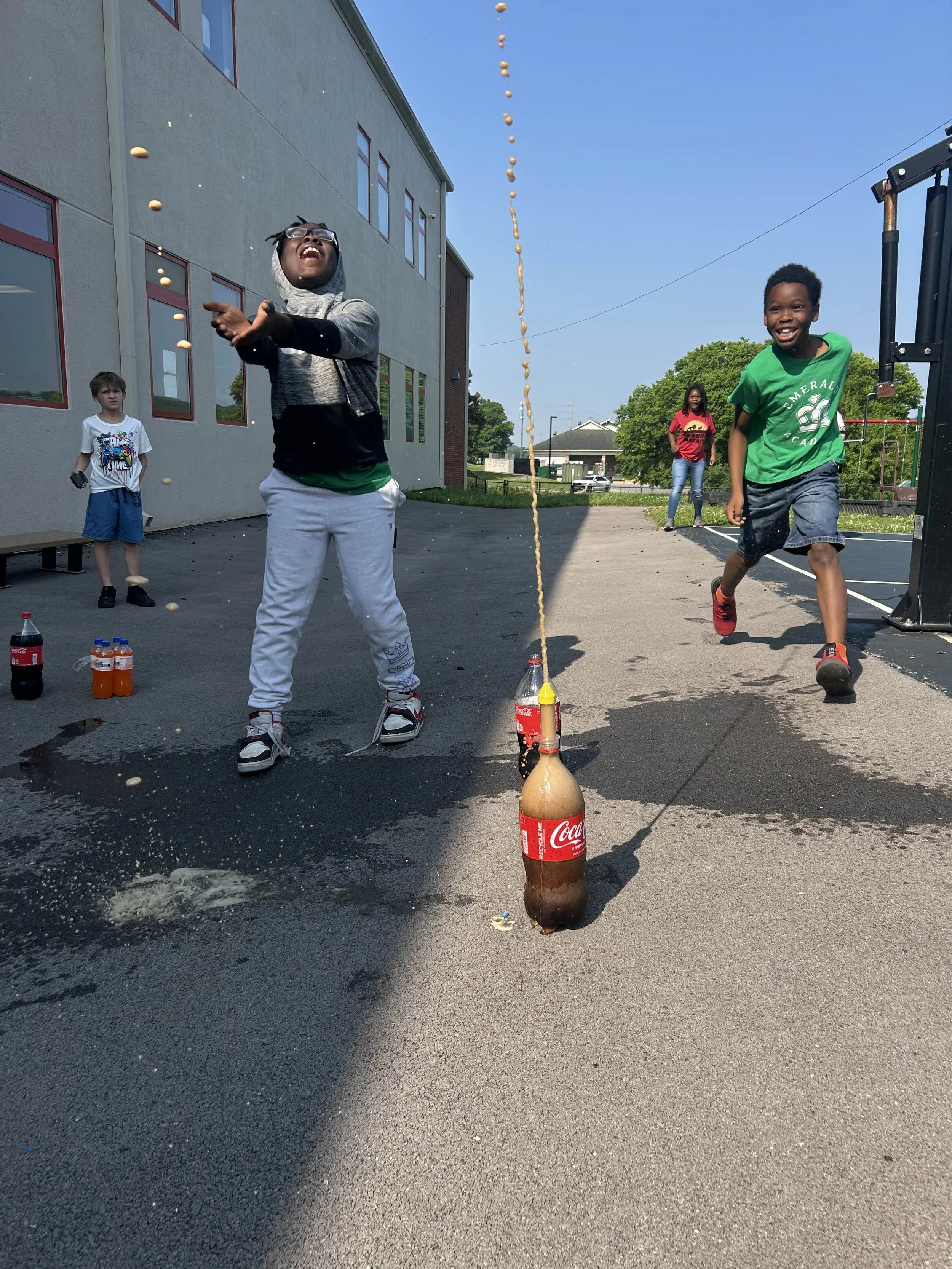 Kids playing and throwing soda bottles into the air outside on playground pavement.