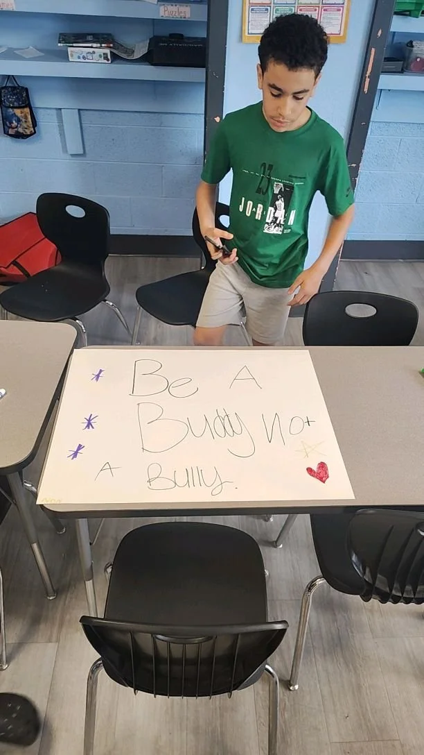 A boy in a green T-shirt and beige shorts standing next to a table with a large white poster that reads 'Be A Buddy Not A Bully' in a classroom setting.