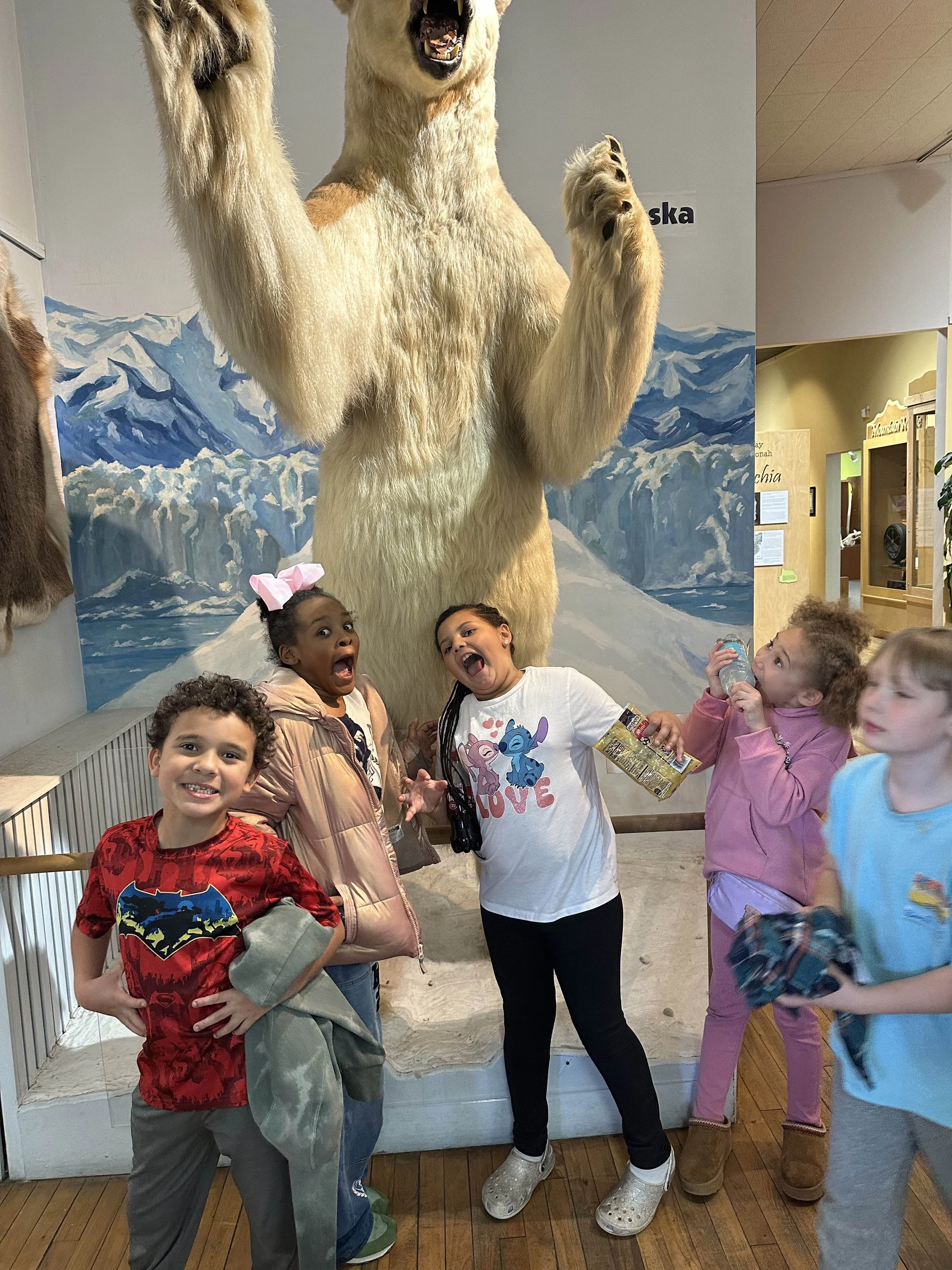 Children at a museum taking a photo with a large model of a bear standing on its hind legs, with mountain scenery in the background.