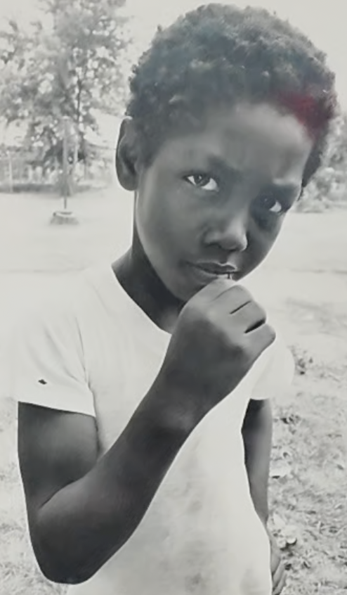 A young boy with short curly hair and dark skin, wearing a white t-shirt, stands outdoors with trees and an open area in the background. He has a serious expression and holds his hand near his mouth.