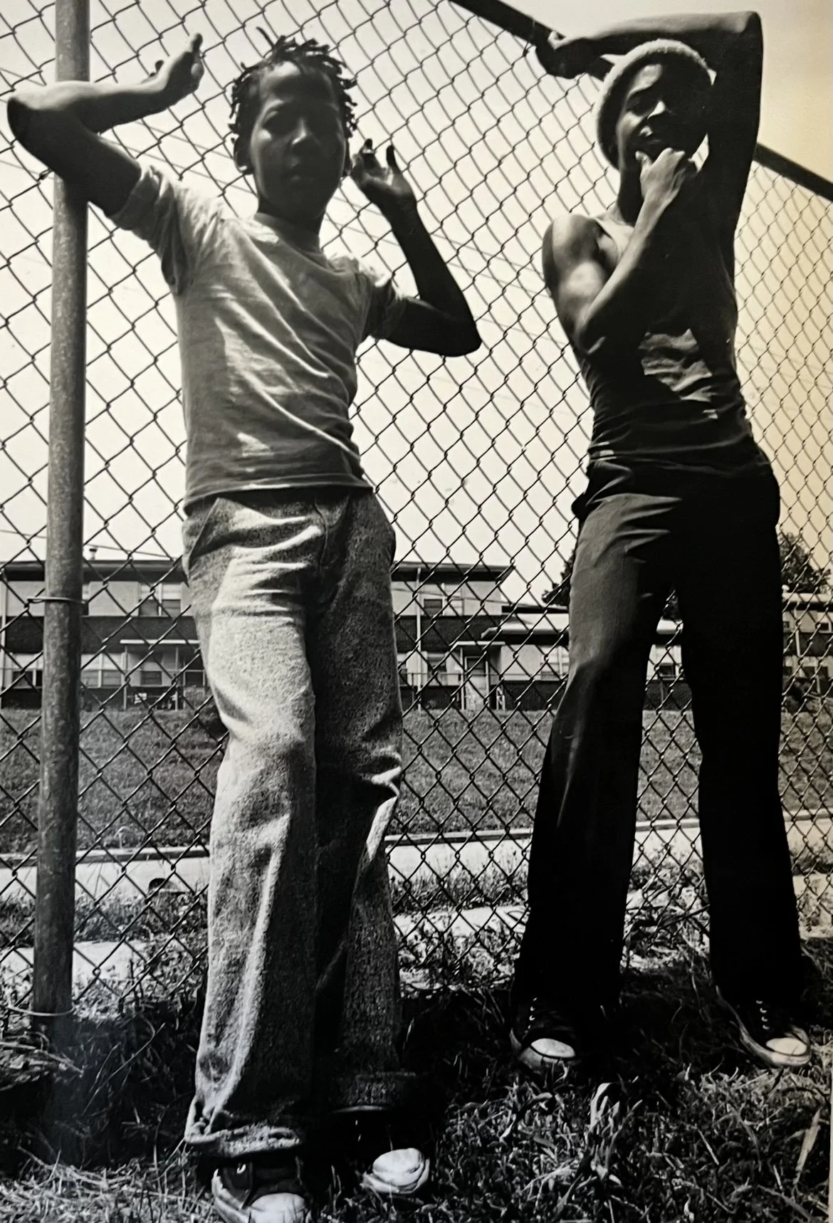 Two young boys standing in front of a chain-link fence, with houses and a yard visible in the background, posing for the photo.