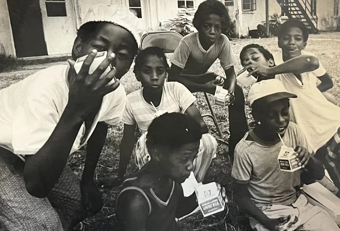 Group of children sitting and standing outside, some drinking from juice boxes, in front of a building with fire escape stairs.