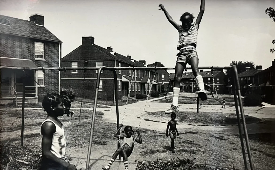 Black children playing on a swing set in a residential neighborhood, with houses and a lawn in the background.