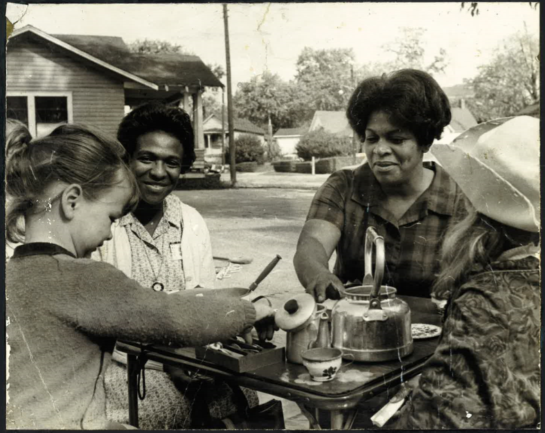 Four women gathered around a small outdoor table with a teapot, cups, and various small items, engaging in a social activity on a residential street.