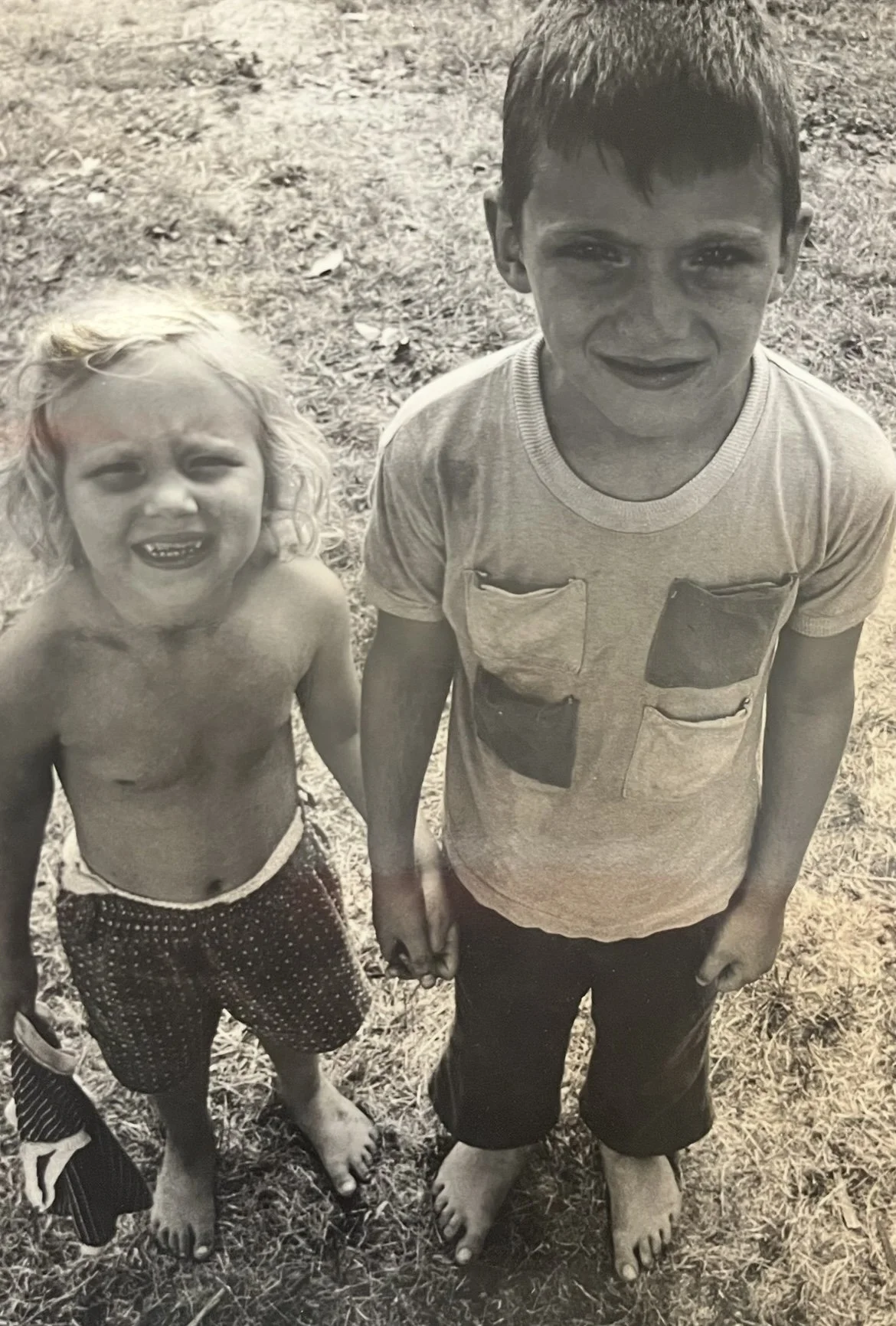 Two children, a girl with curly hair and a boy, are standing outdoors and holding hands, both barefoot, with a dirt ground and grass in the background.