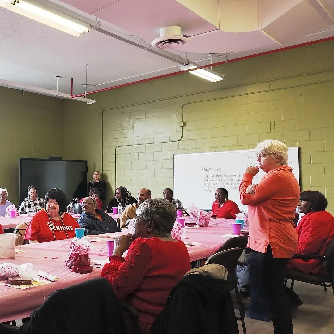 A group of diverse seniors sitting around pink-covered tables in a community room, listening to a woman in an orange sweater speaking, with a whiteboard and large monitor in the background.