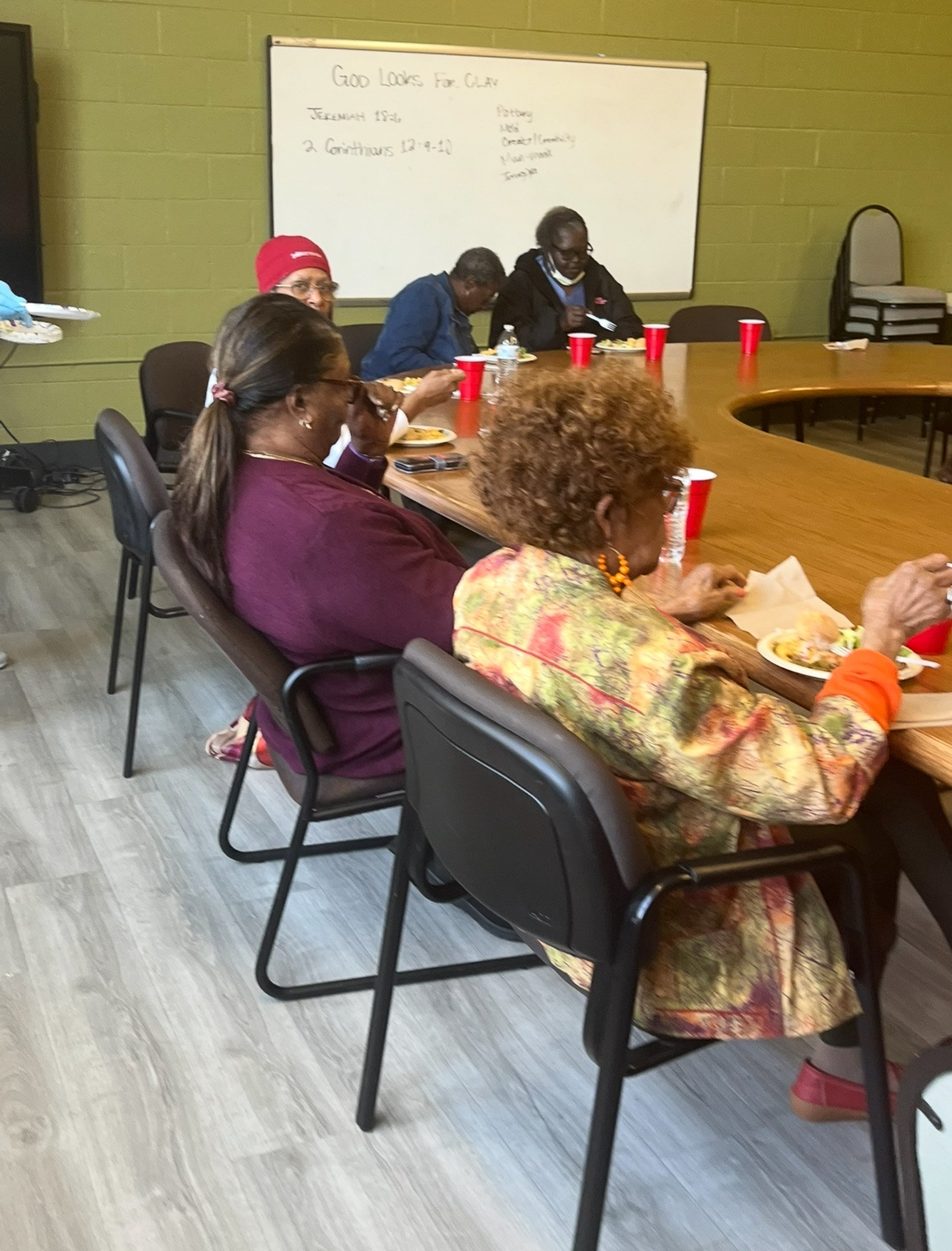 A group of people sitting around a large, curved wooden conference table eating a meal. There are red cups and plates on the table. A whiteboard with writing is in the background.