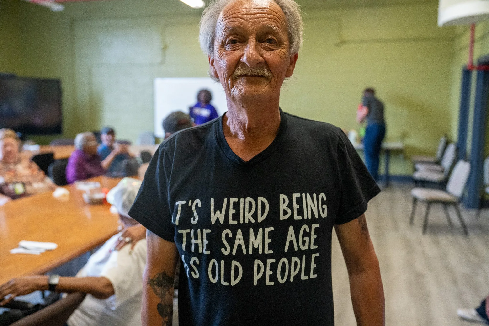 An elderly man with gray hair and a mustache smiling at the camera, wearing a black T-shirt that reads 'It's weird being the same age as old people', in a room with other people seated at tables in the background.