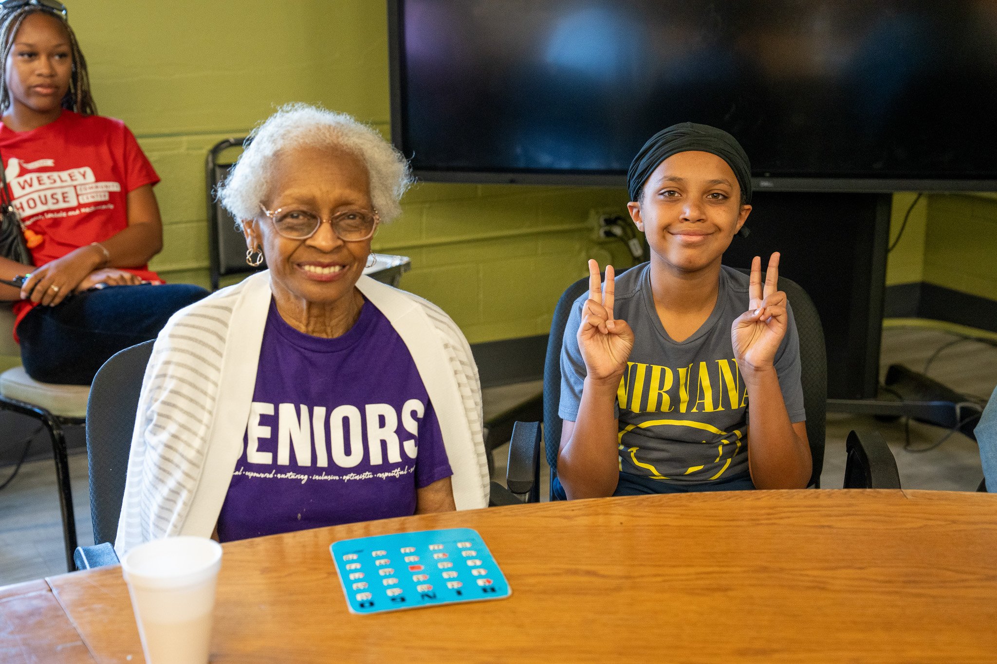 An elderly woman with white curly hair and glasses sitting at a table, wearing a purple t-shirt with 'SENIORS' printed on it, and a young boy wearing a grey Nirvana t-shirt and black head wrap showing peace signs with both hands, smiling. There is a 