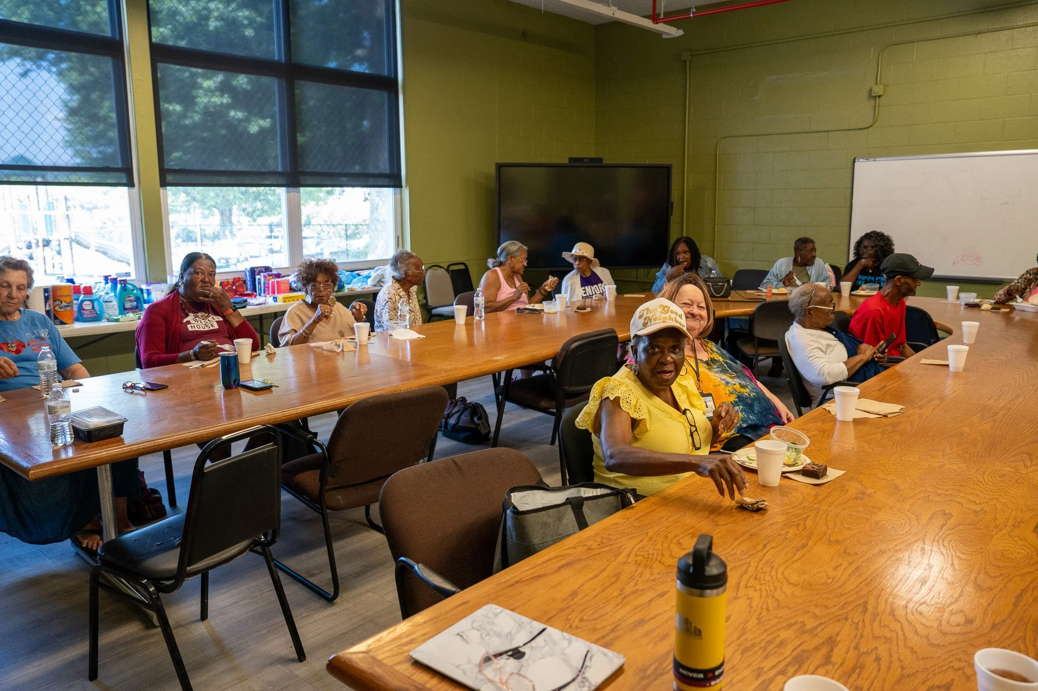 A group of diverse women gathered in a meeting room, sitting around large wooden tables. Some are eating, drinking, or engaging in conversation. The room has large windows with blinds, a whiteboard, and a large screen on the wall. Refreshments and su