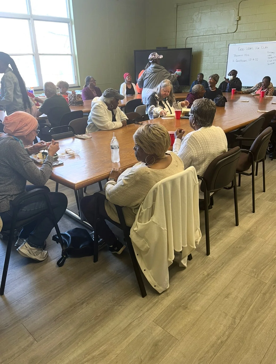 Group of older adults sitting around a large rectangular conference table in a well-lit room, some eating and some engaging in conversation, with a large screen and a whiteboard in the background.