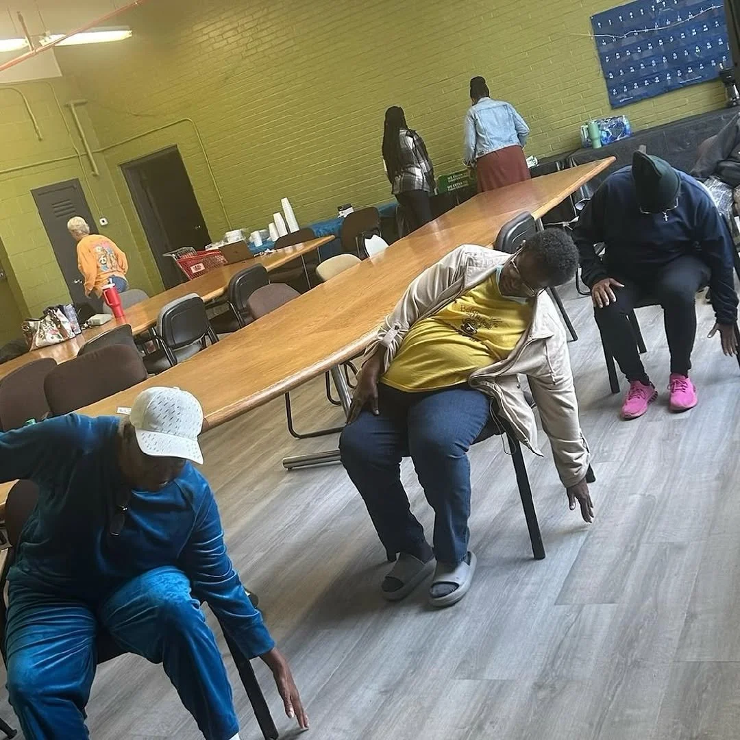 People participating in a stretching or exercise activity in a room with long tables, chairs, and a brick wall.