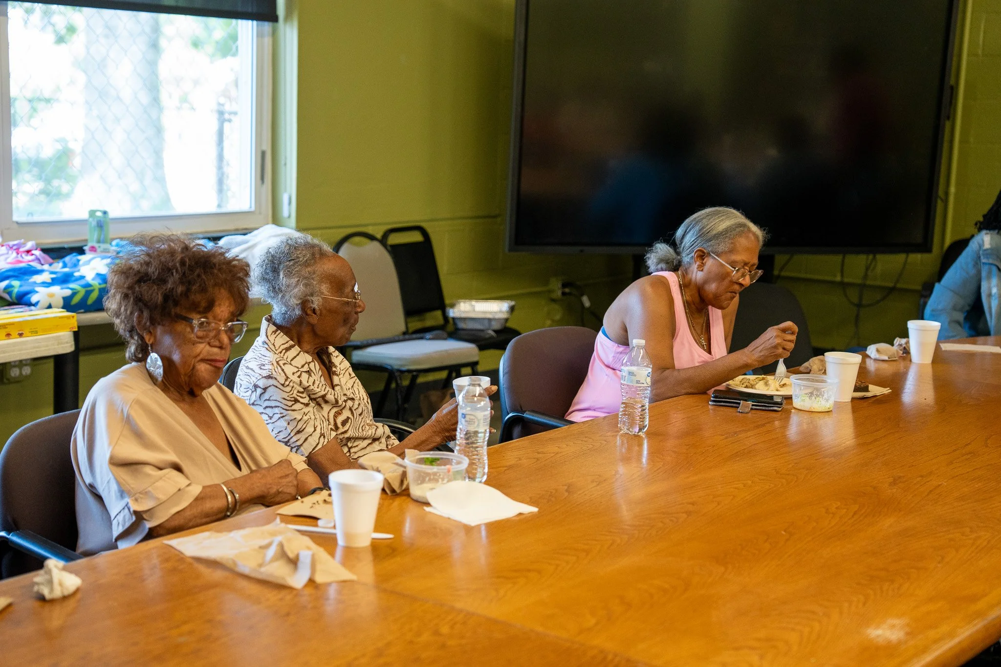 Four elderly women sitting at a wooden table eating and drinking in a room with green walls and large windows.